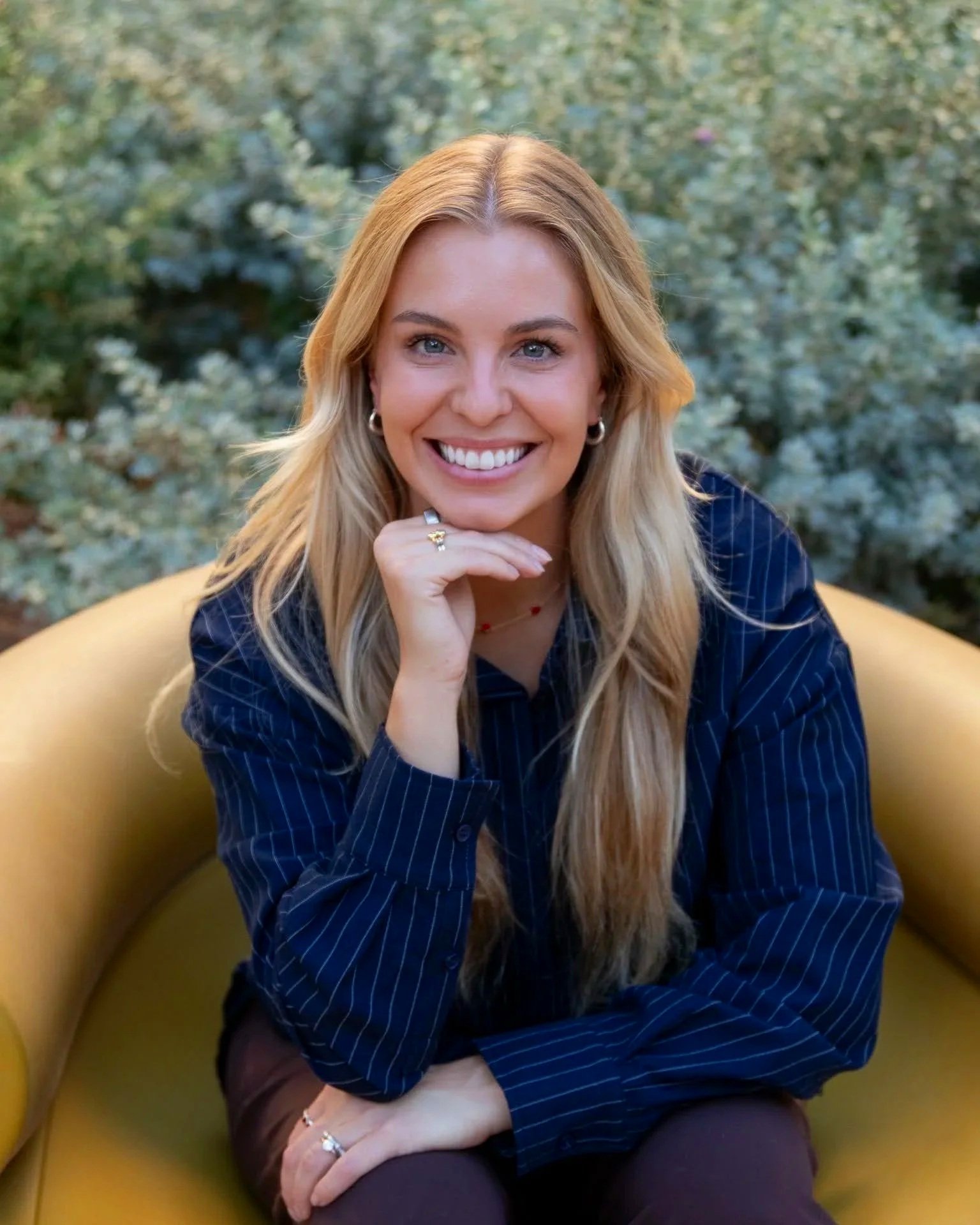 A smiling woman with long blonde hair sitting on a yellow couch outdoors, with a background of green plants.