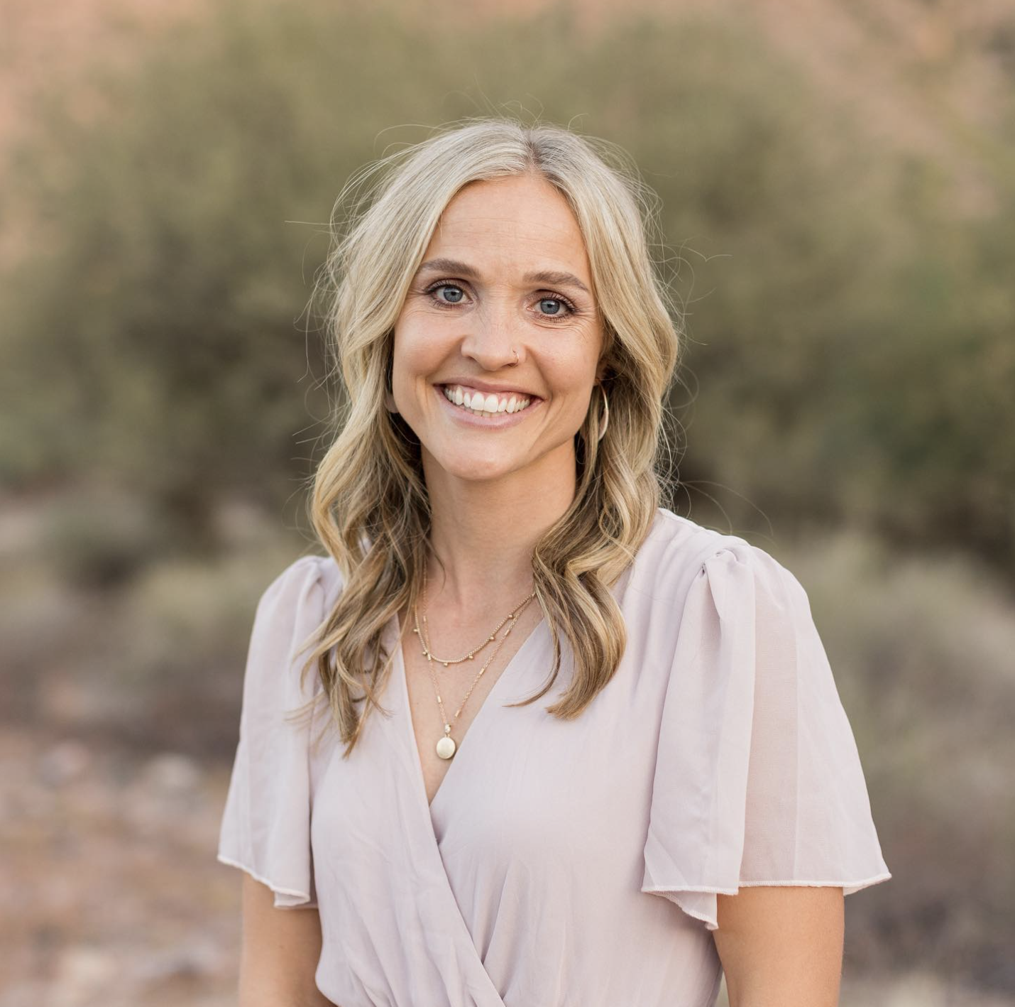 A smiling woman with blonde hair, wearing a light beige blouse and layered necklaces, outdoors with a blurred natural background.
