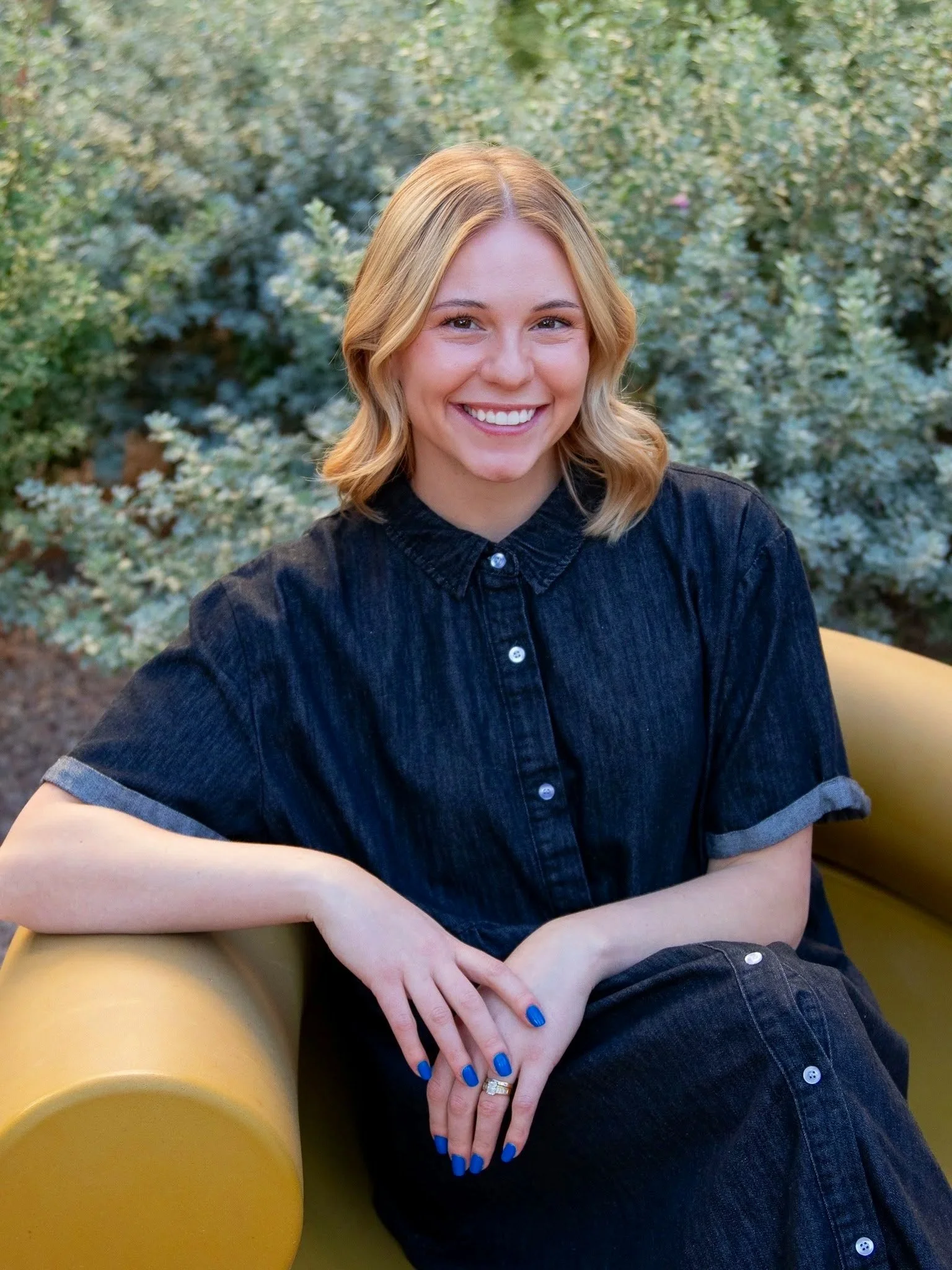 A young woman with blond hair and a black denim dress, sitting on a yellow bench outdoors, smiling at the camera with greenery in the background.