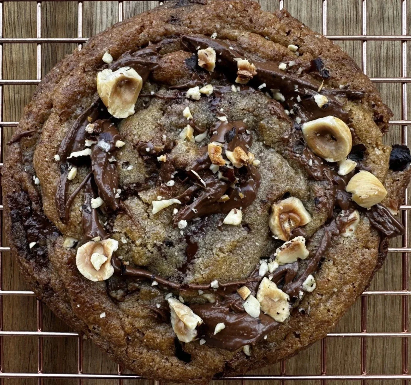 A large chocolate chip cookie topped with chocolate drizzle and chopped nuts, resting on a cooling rack.