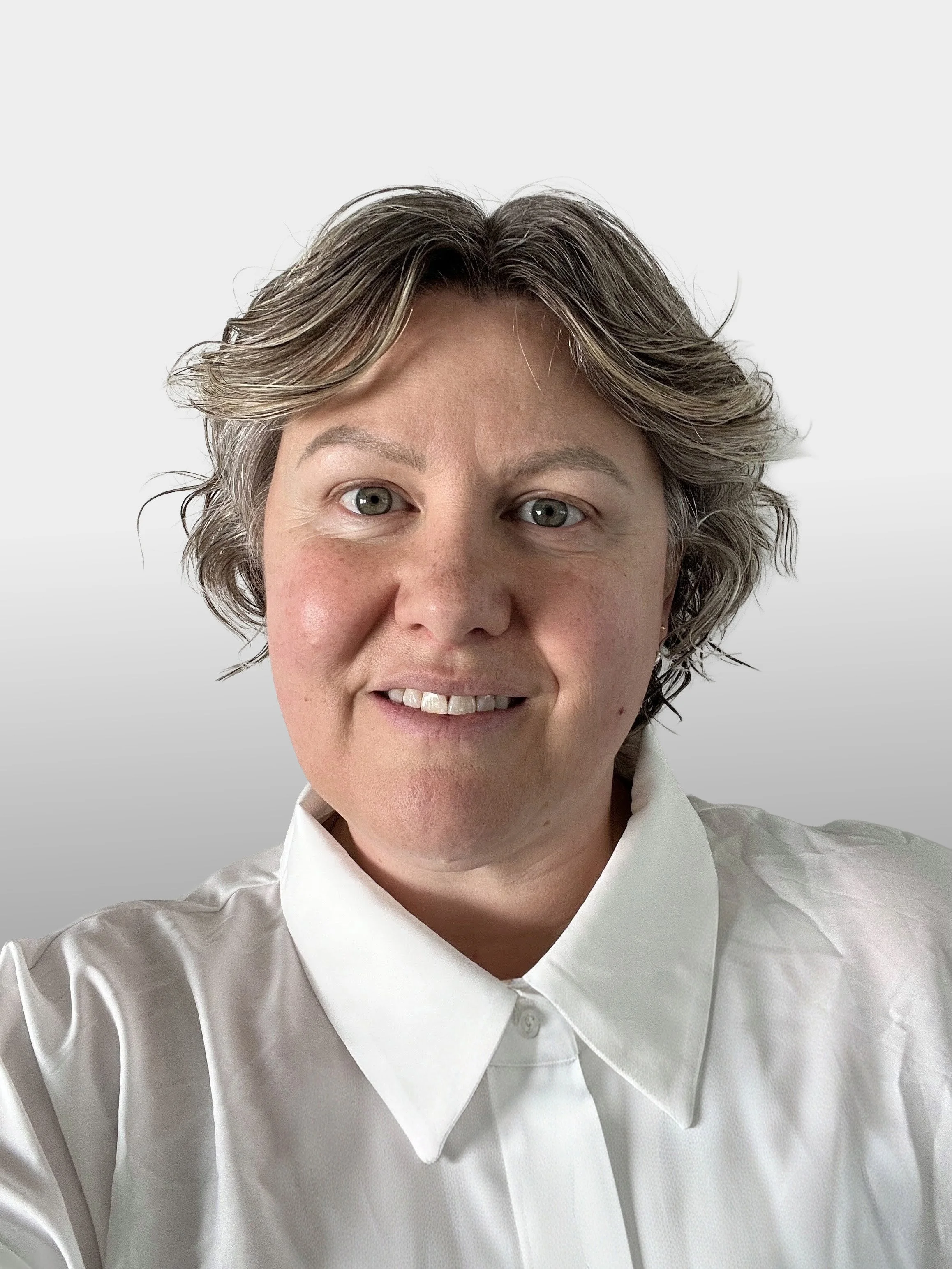 A person with short, wavy, gray hair, light skin, and wearing a white collared shirt, smiling in a close-up photo.