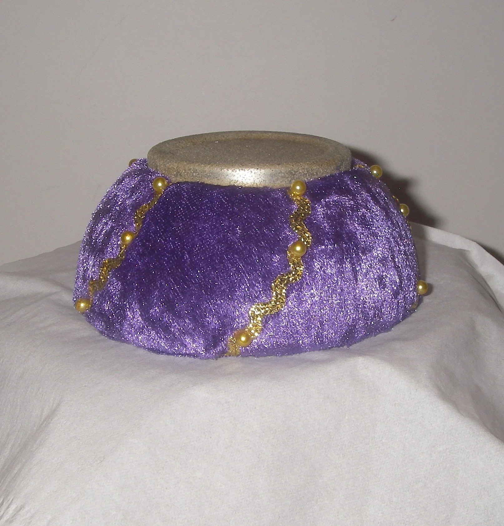 Decorative purple and gold ornament with glitter, beads, and a silver top, resting on white fabric