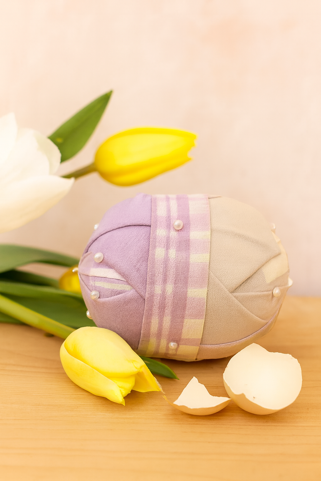A fabric egg decorated with pearls, surrounded by yellow tulips and white eggshells on a wooden surface.