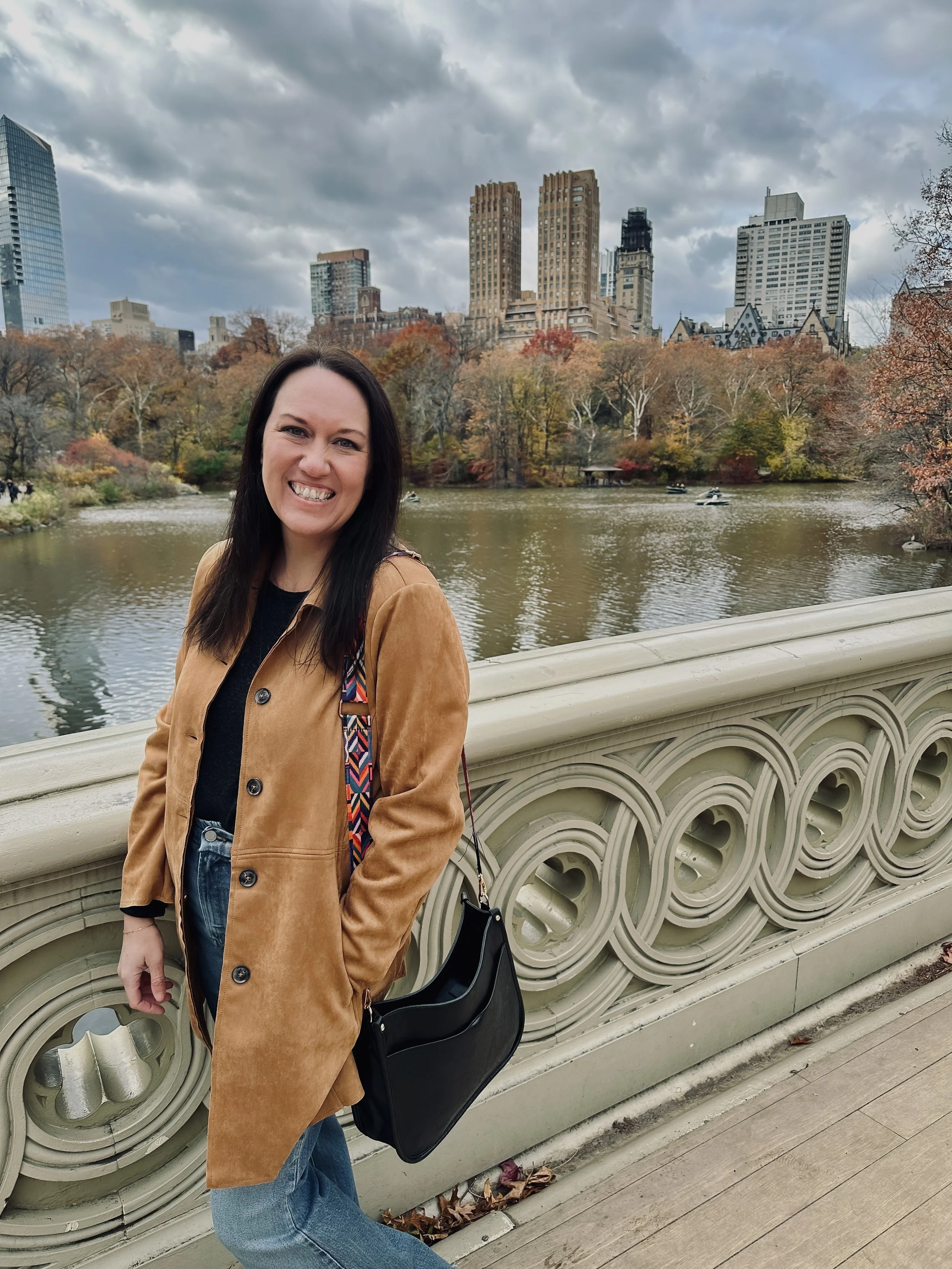 Woman smiling on a bridge over a lake in Central Park with city skyscrapers in the background during autumn.