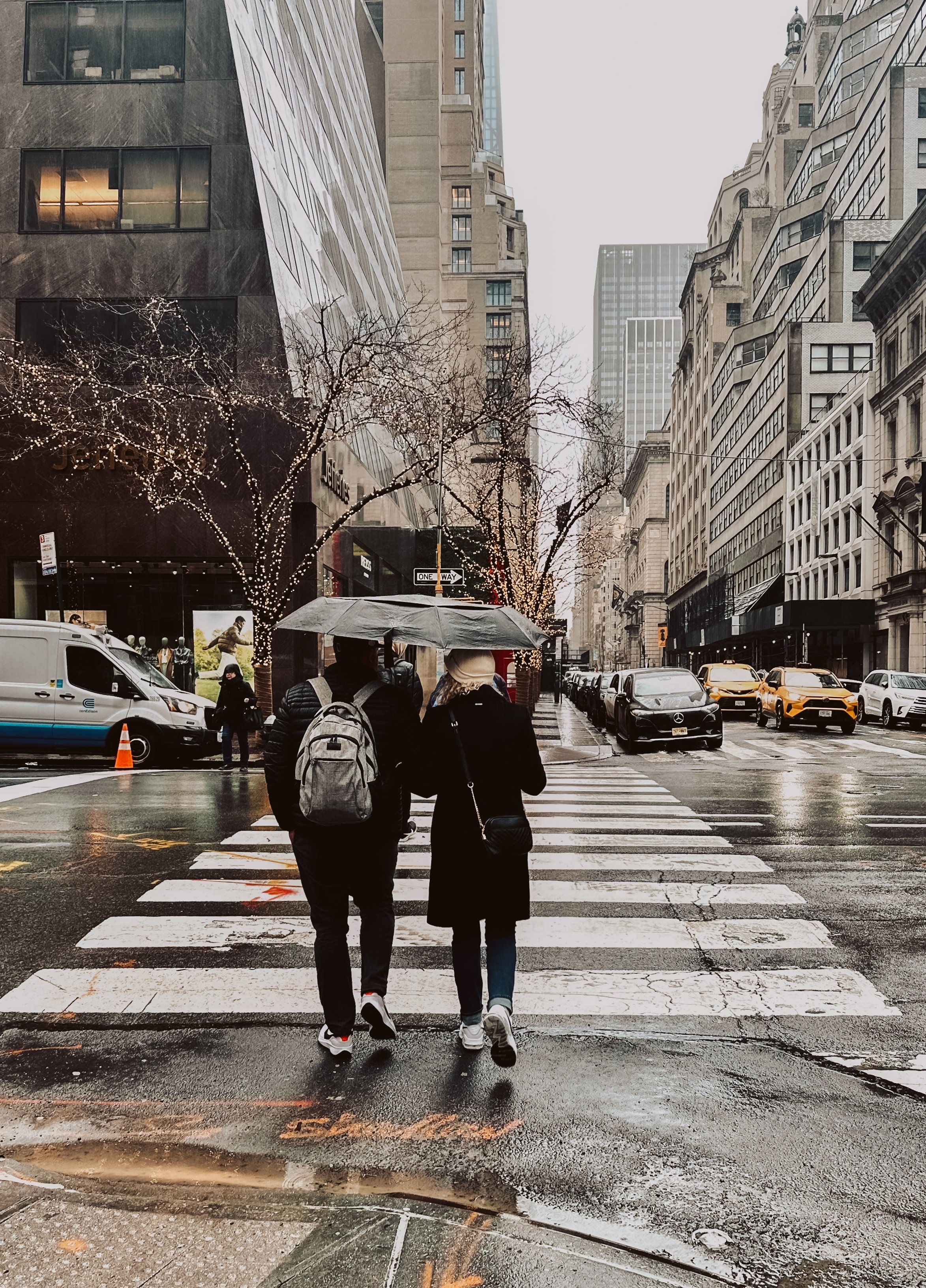 Two people walking together on a crosswalk in an urban city with tall buildings, some of which are decorated with string lights, on a rainy day holding an umbrella.
