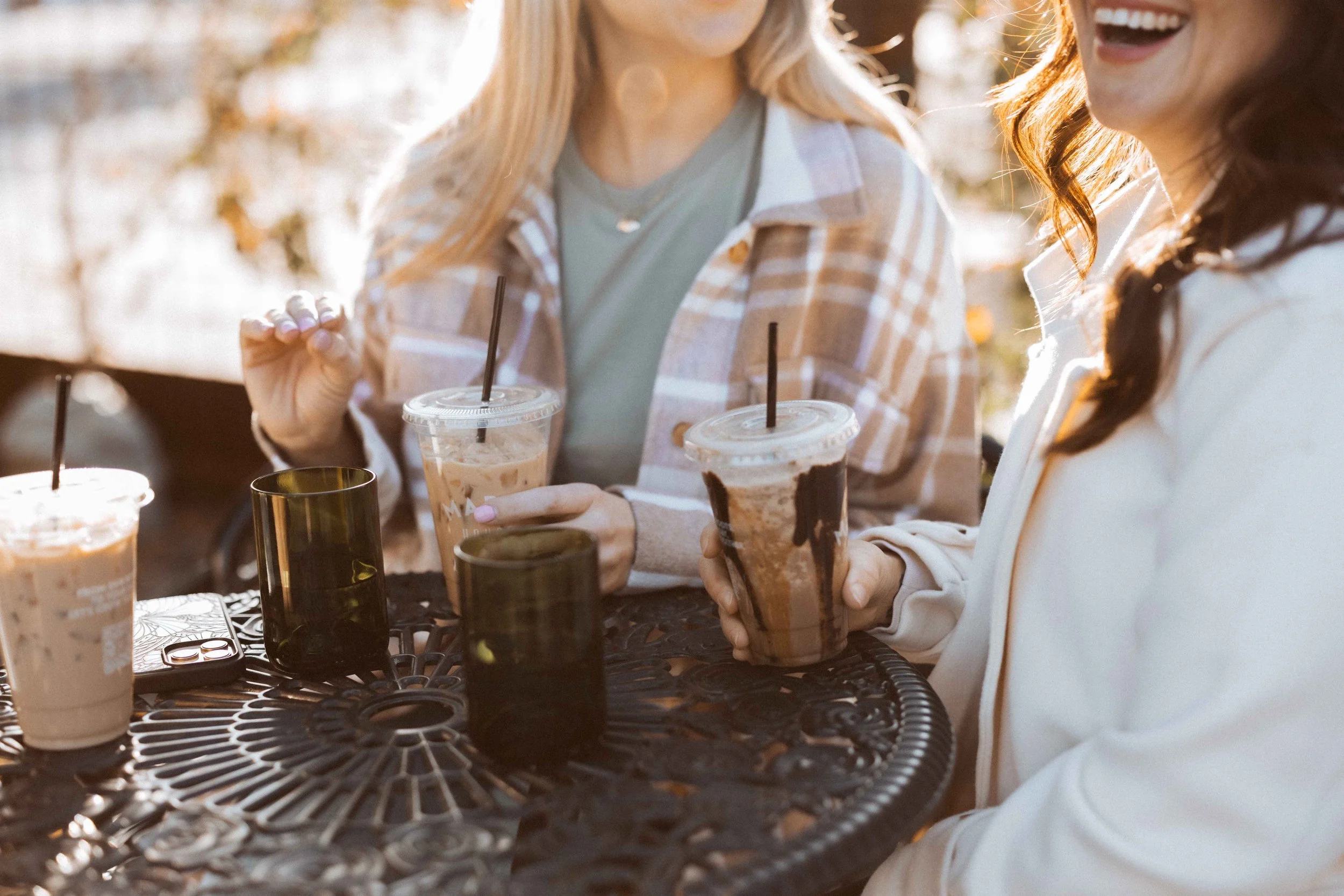 Two women are sitting at an outdoor table with five iced coffee drinks and two black glasses on it, enjoying a sunny day.