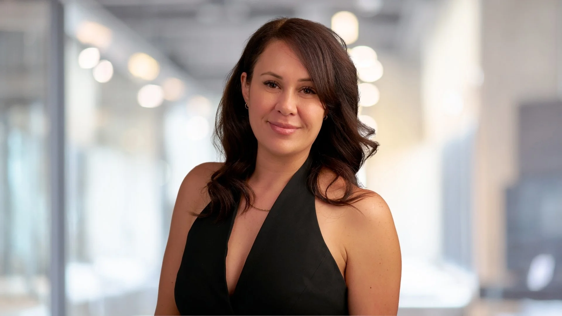 A woman with shoulder-length dark brown hair and light skin, smiling while wearing a black sleeveless top, standing in a modern, brightly lit office or indoor space.