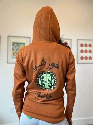 Image of a young woman wearing the orange "Khorshid Khanoom" hoody against a backdrop of a couple of pieces from the exhibit.