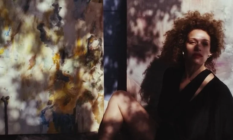 Image of artist Sita Zimmer facing the camera and sitting in front of two of her pieces.