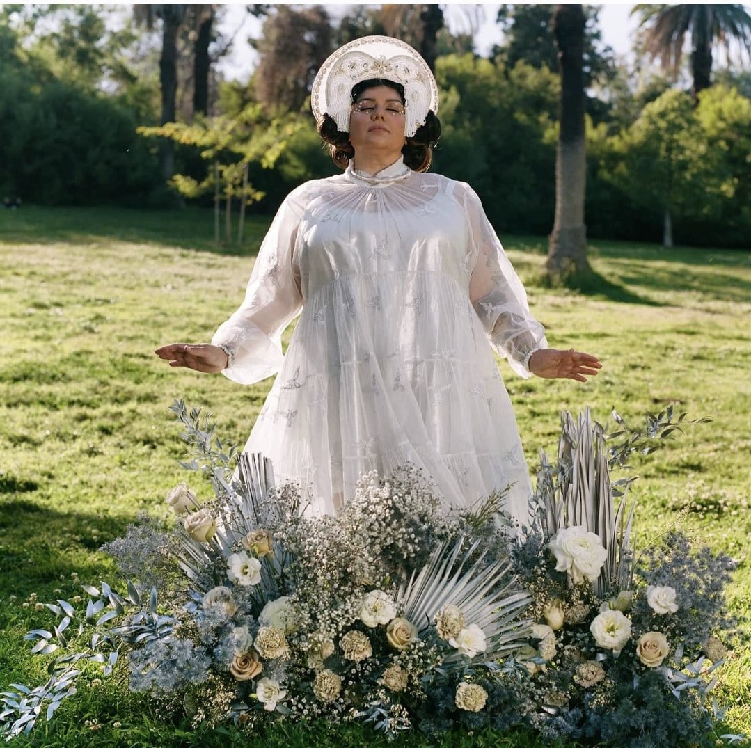 Image of artist Ever Velasquez dressed in a white gown in front of ceremonial flowers surrounded by grass and trees.