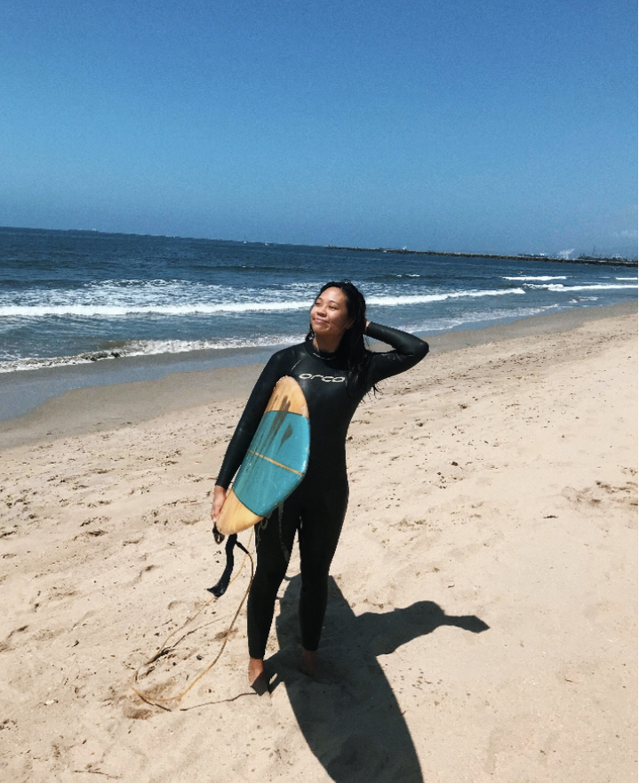 Image of artist Gabrielle Chen in a wetsuit holding a surfboard at the beach.