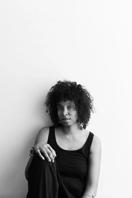 Black and white image of artist ThaooAishat Hasati sitting against a plain wall facing the camera.