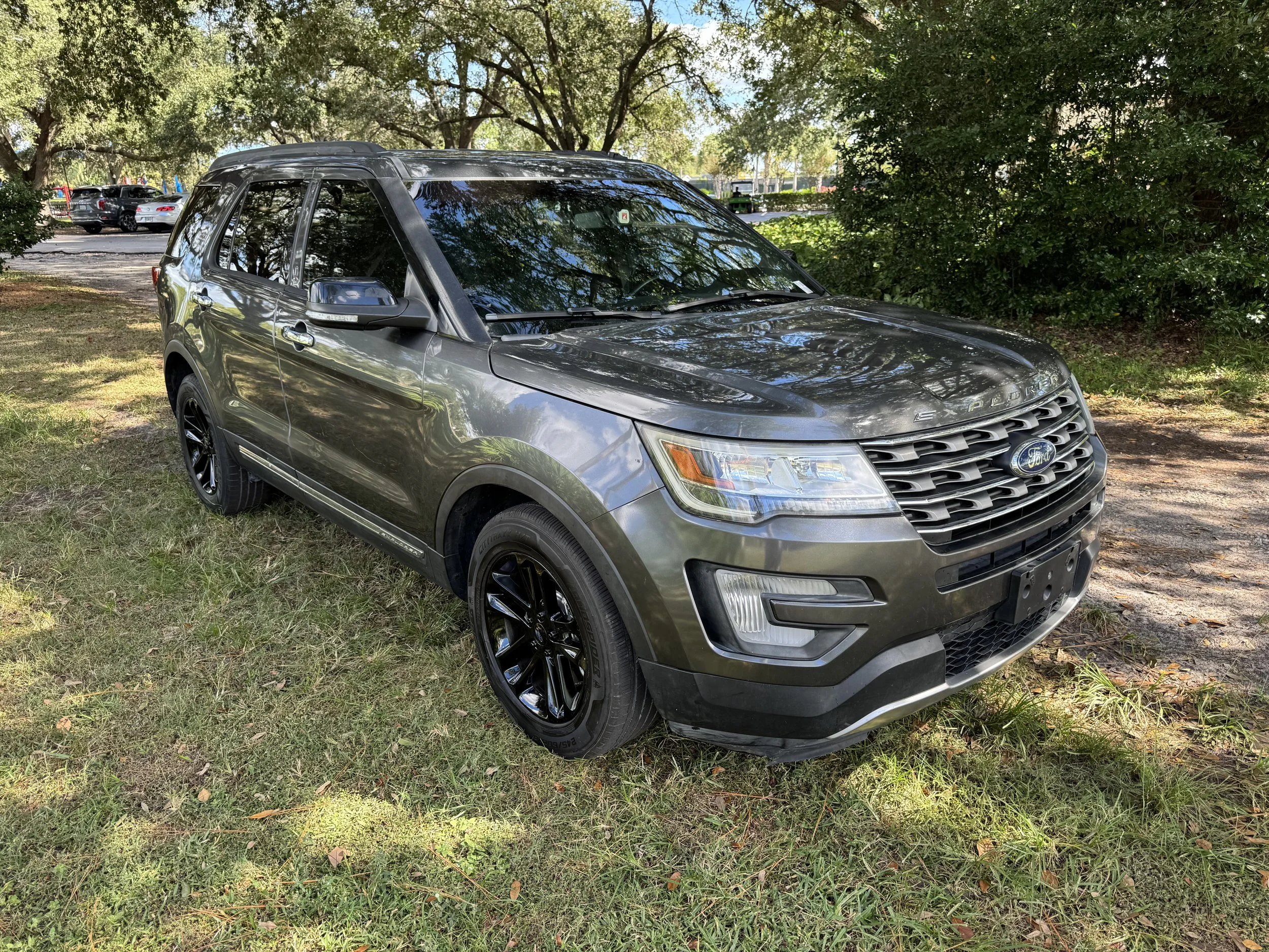 Gray Ford Explorer SUV parked on grassy area under trees, with a parking lot and other cars in background.