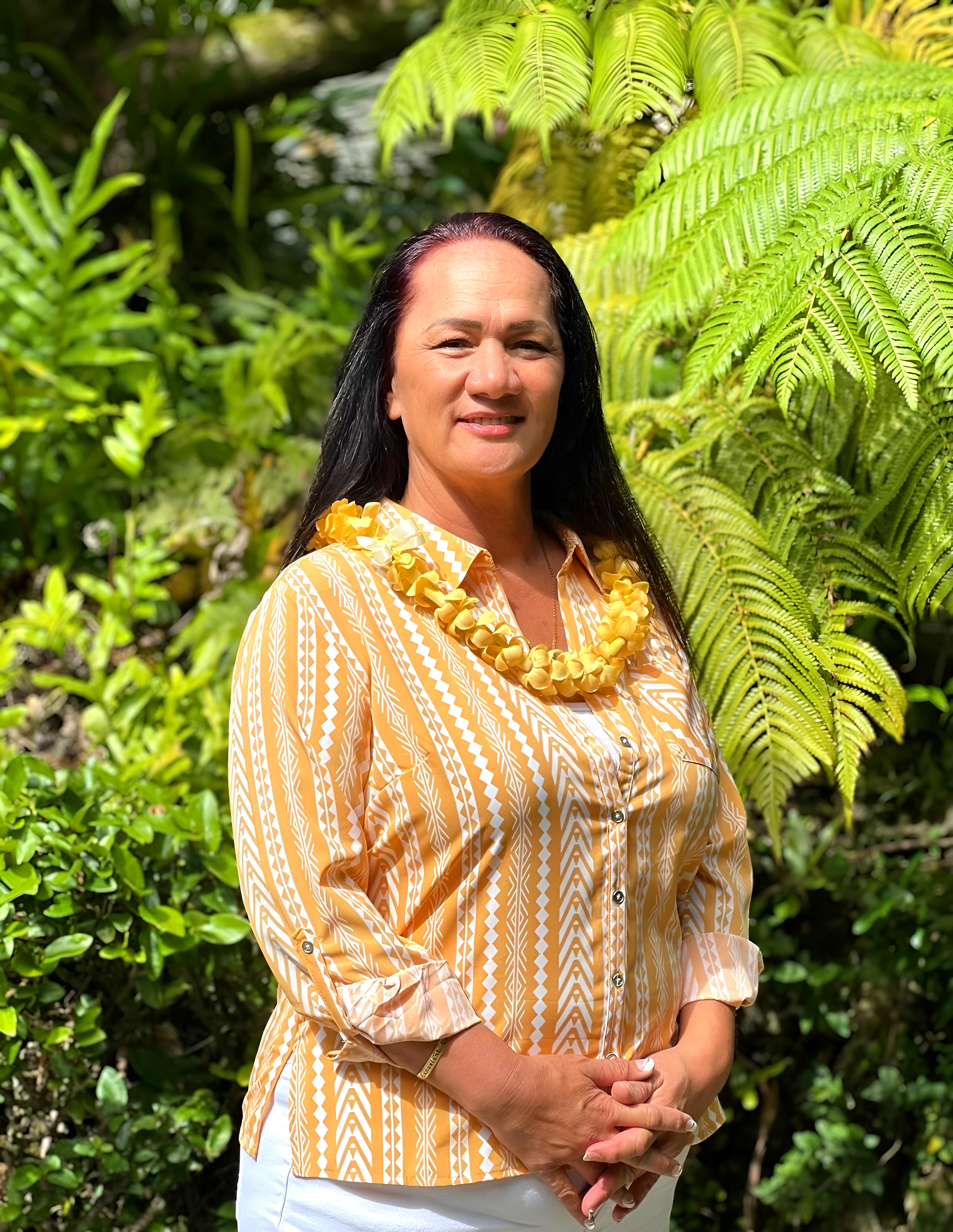 A woman with long dark hair, wearing a yellow patterned blouse and a yellow floral necklace, stands outdoors in front of lush green tropical plants.