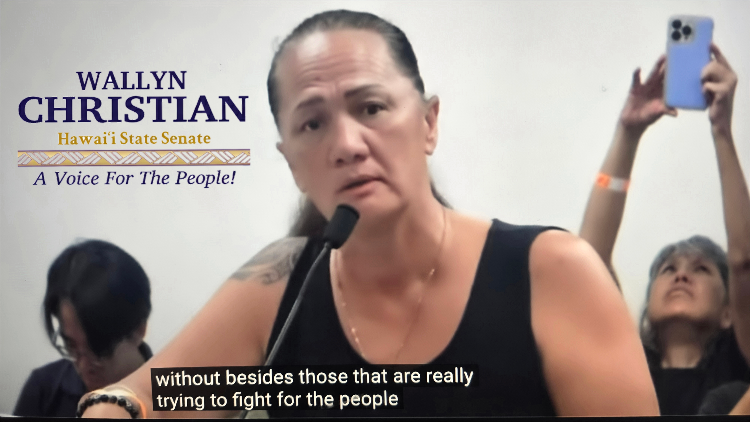A woman at a public meeting or event speaking into a microphone with a serious expression. Behind her, a person is taking a photo with a smartphone, and another individual is sitting and looking down. There is a sign on the wall that reads 'WALLYN CHRISTIAN Hawaii State Senate A Voice For The People!'.