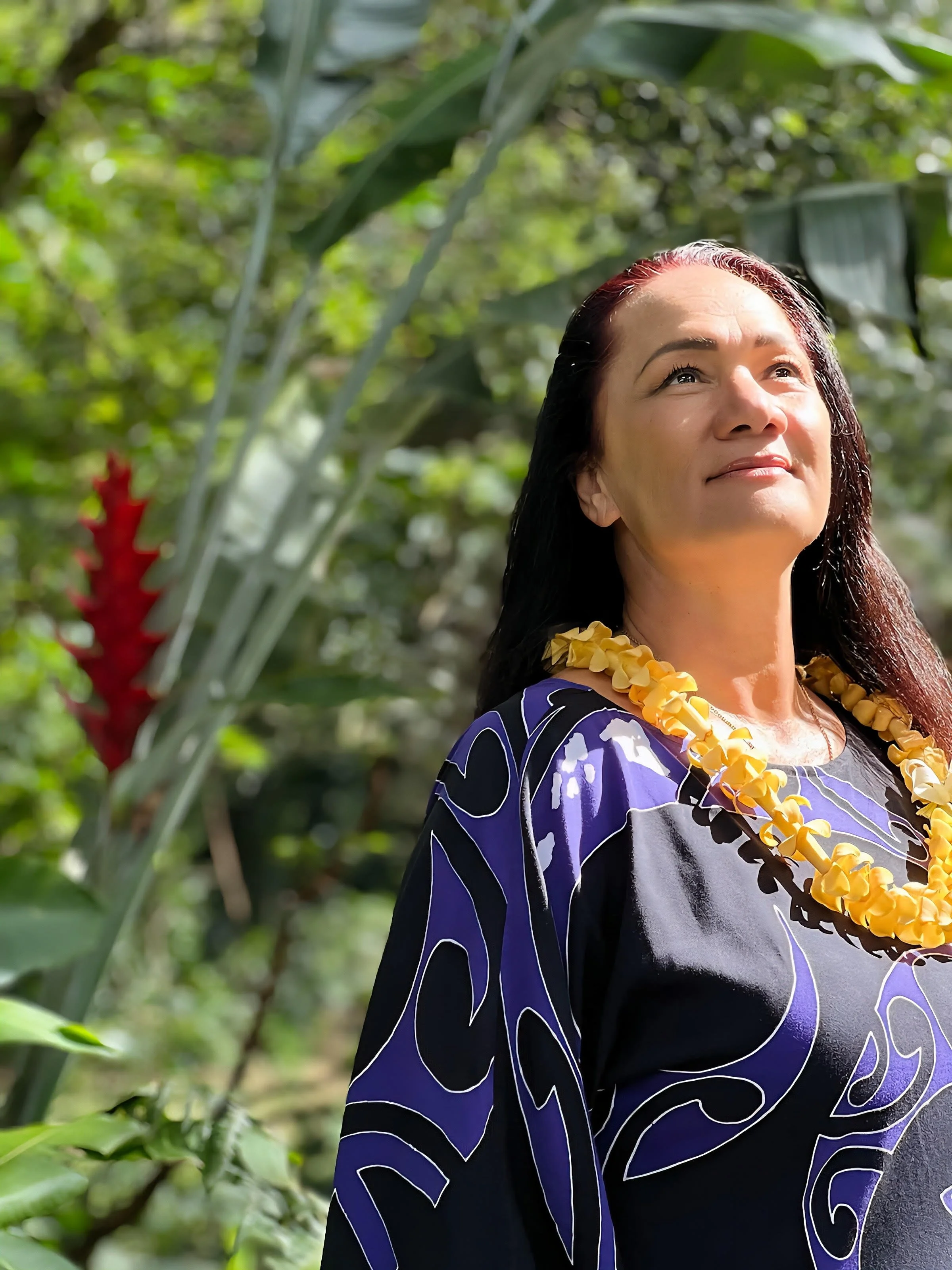 A woman with dark hair and a fair complexion stands outdoors among tropical greenery, wearing a purple and black patterned dress and a yellow flower lei, looking up with a serene expression.