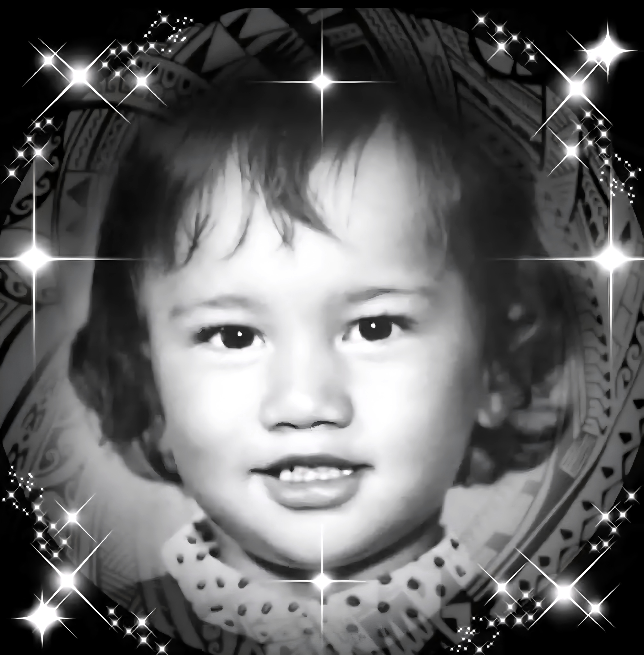 Black-and-white photo of a young child with curly hair, smiling, wearing a polka dot shirt, surrounded by decorative sparkles and a patterned background.