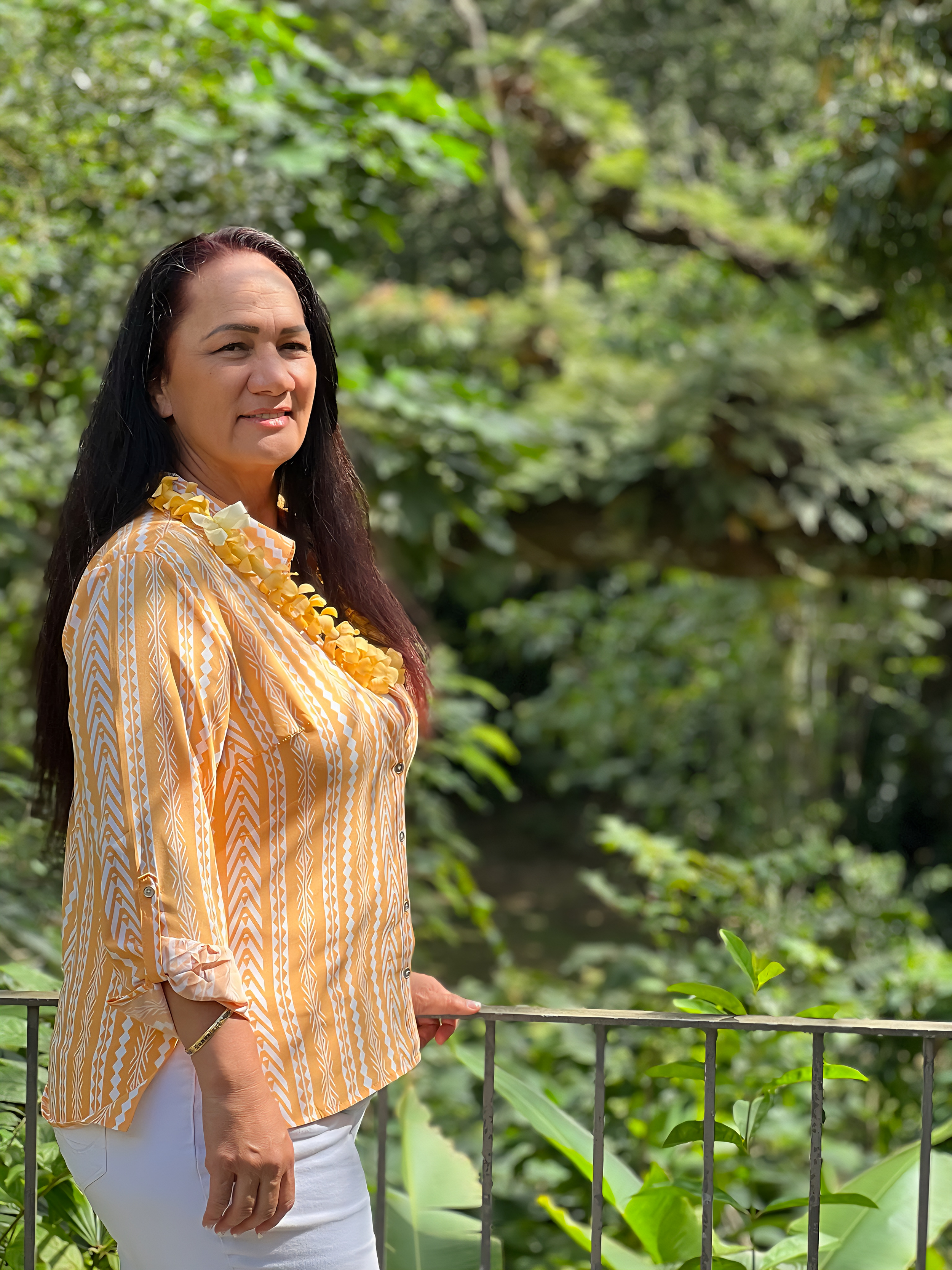 Wallyn Christian standing outdoors on a balcony, wearing a yellow and white patterned blouse and a flower lei, with lush green foliage in the background.