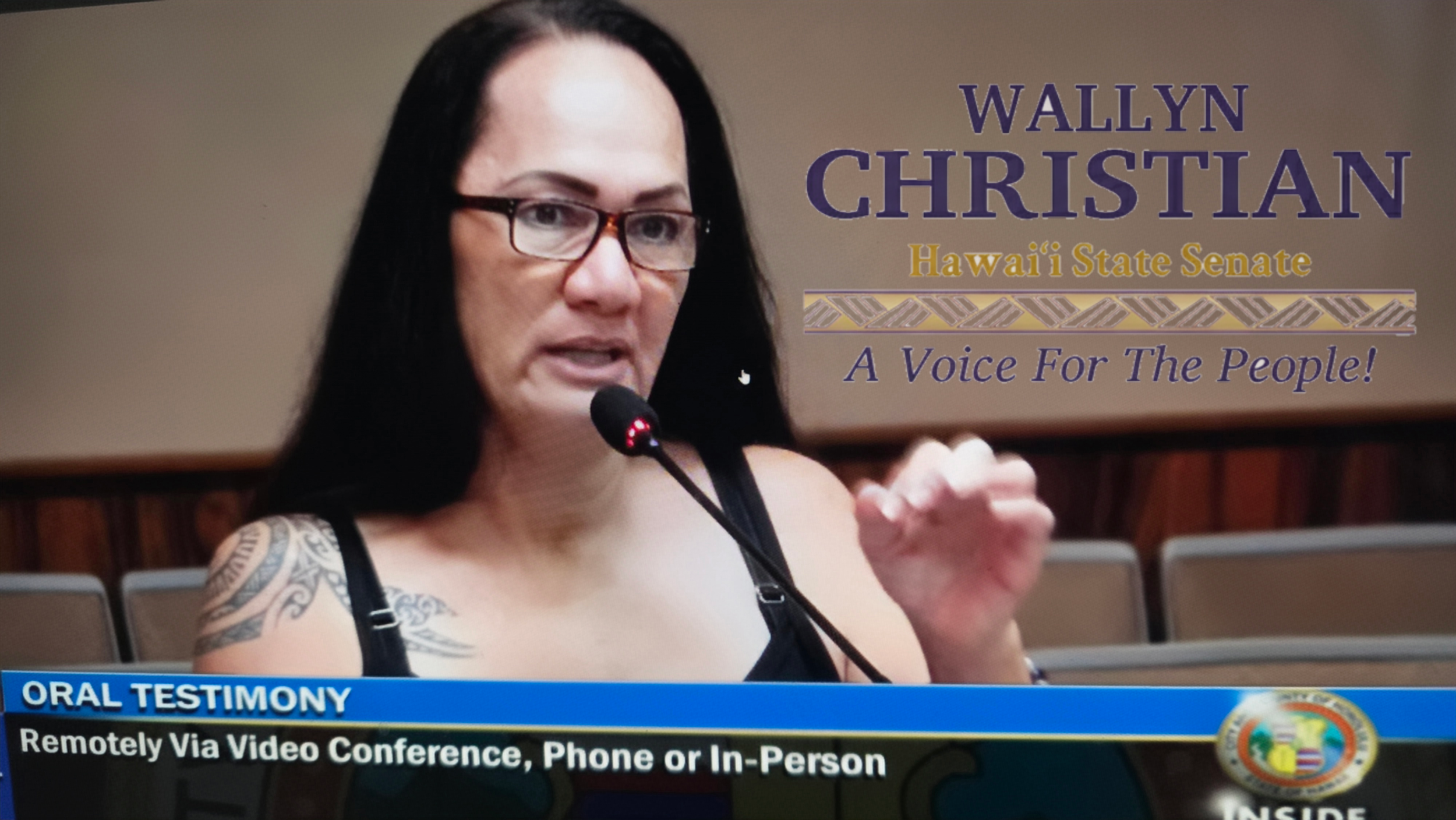A woman with long dark hair and glasses speaking into a microphone during a televised hearing or testimony. The background contains a banner with her name Wallyn Christian, her title Hawaii State Senate, and slogan 'A Voice For The People'. A blue news overlay at the bottom reads 'ORAL TESTIMONY Remotely Via Video Conference, Phone or In-Person'.