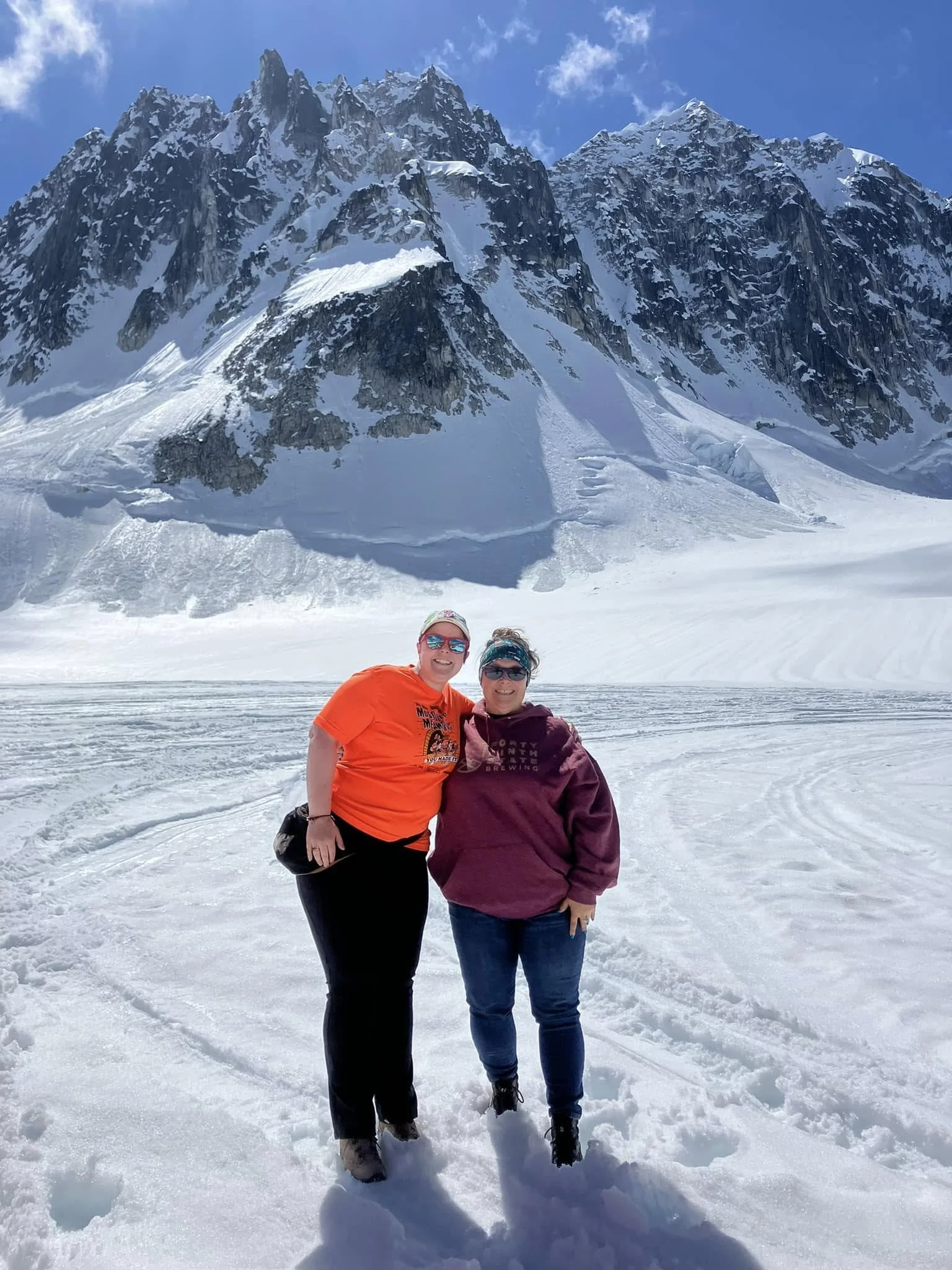 Two people standing on a glacier in front of a towering snow-covered mountain with clear blue sky.