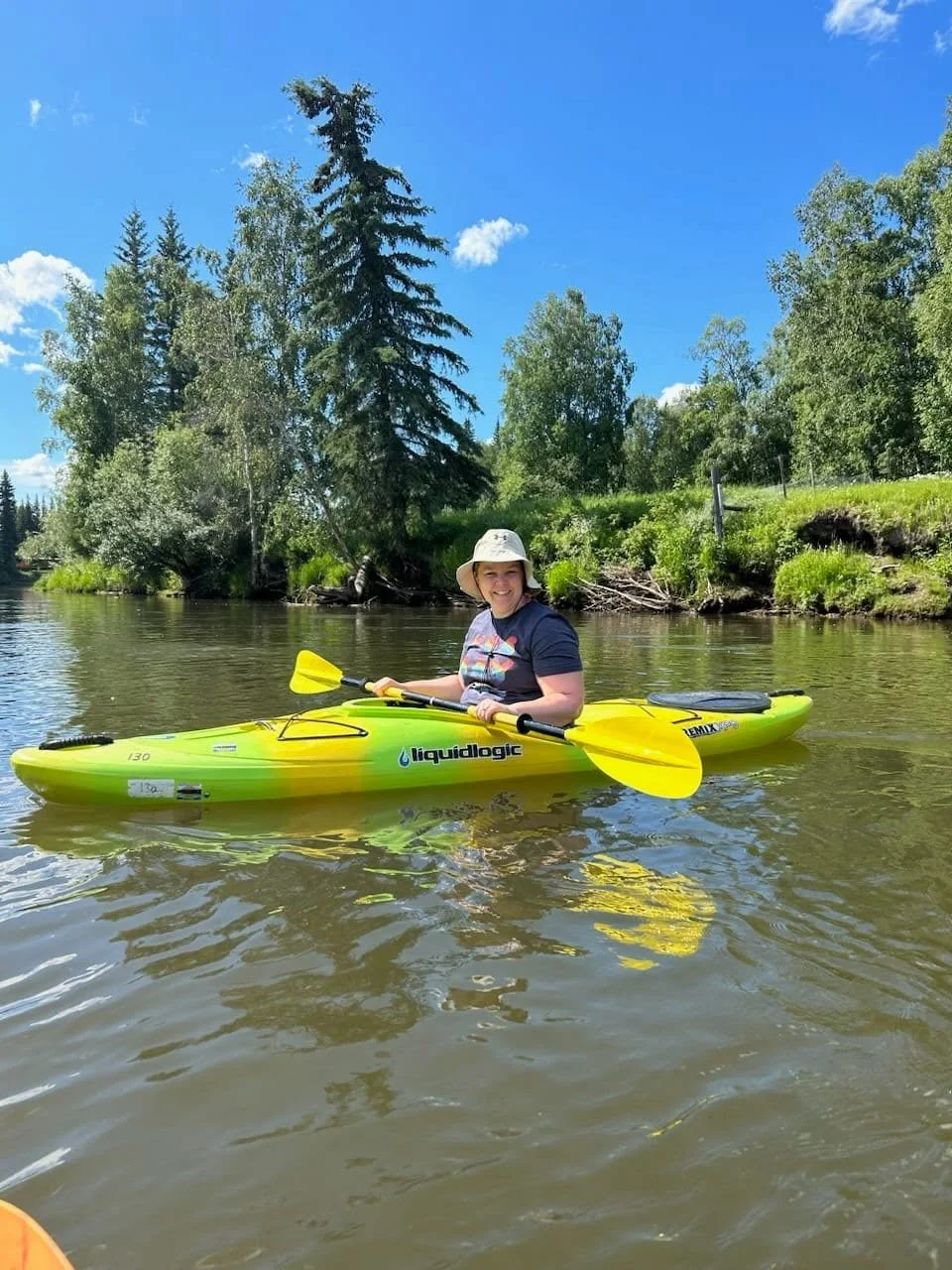 A woman kayaking on a calm river surrounded by trees with a bright blue sky above.