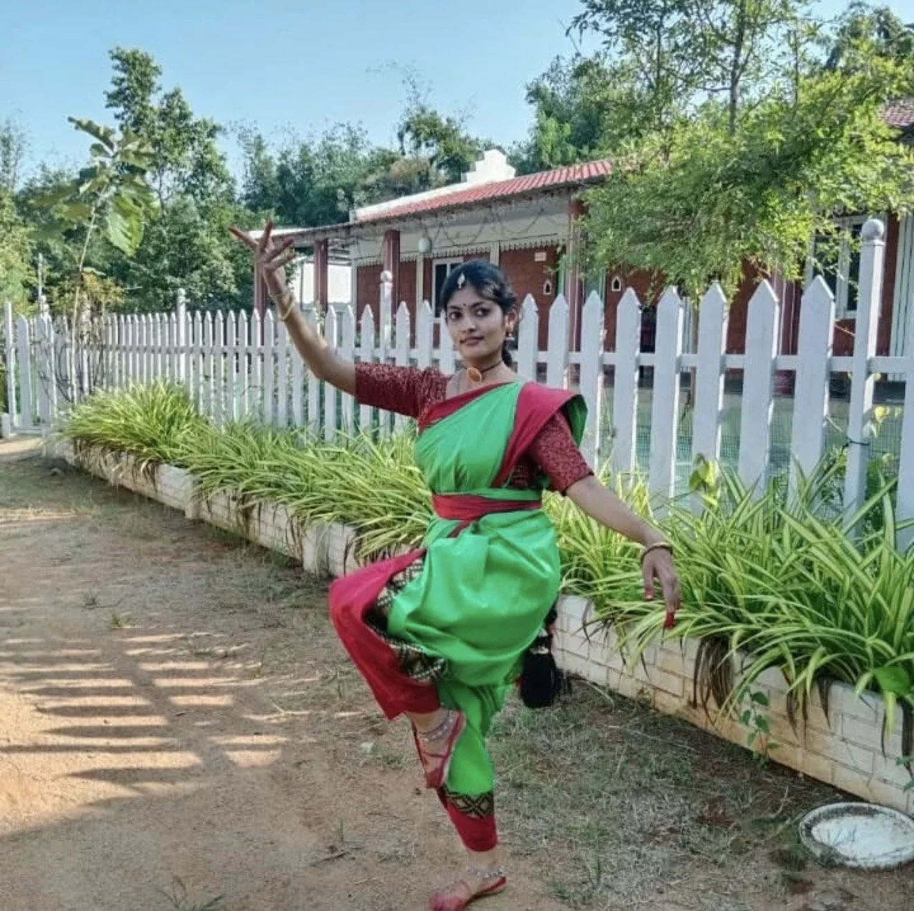A woman wearing traditional Indian attire, a green and red sari, is dancing outdoors on a dirt path by a white fence with greenery and a brick house in the background.