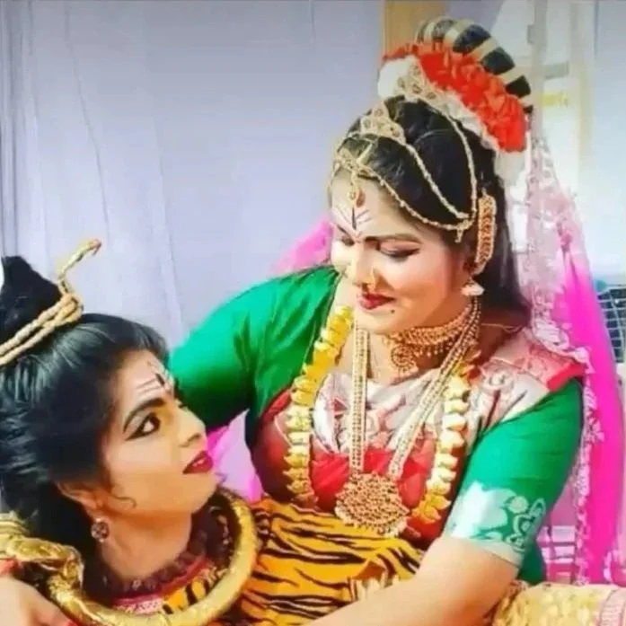 Two women dressed in traditional Indian attire, jewelry, and makeup, with one woman appearing to perform a dance pose while the other looks on.