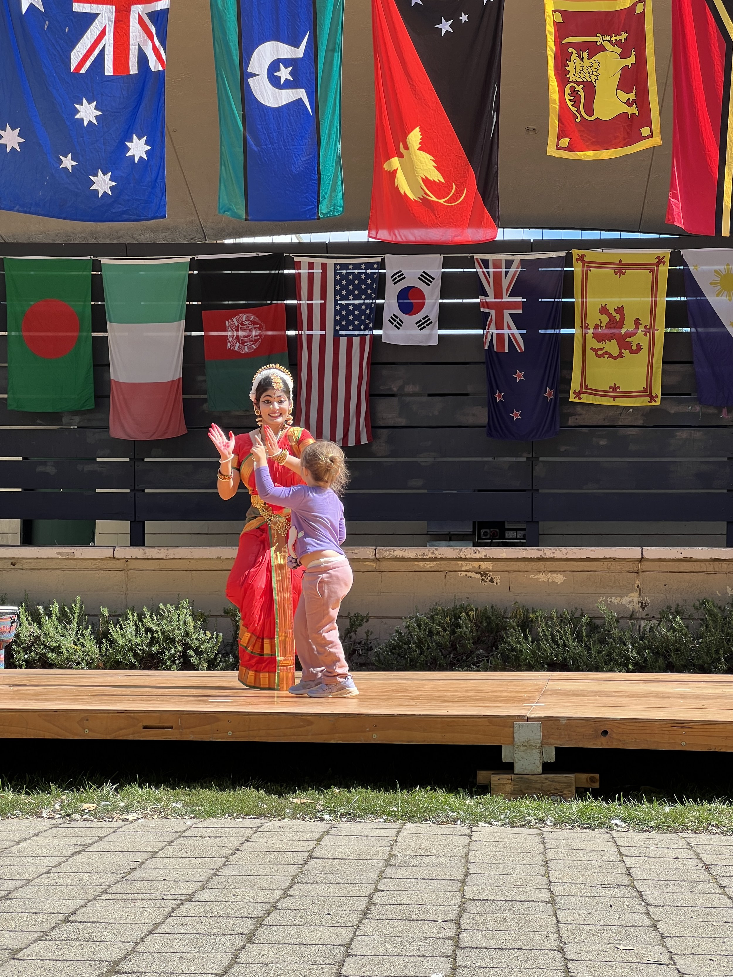 A woman dressed in traditional Indian attire performing a dance on a wooden stage, with a little girl in casual clothing dancing nearby. Behind them, a black fence displays multiple international flags including Australia, Italy, Afghanistan, the Uni