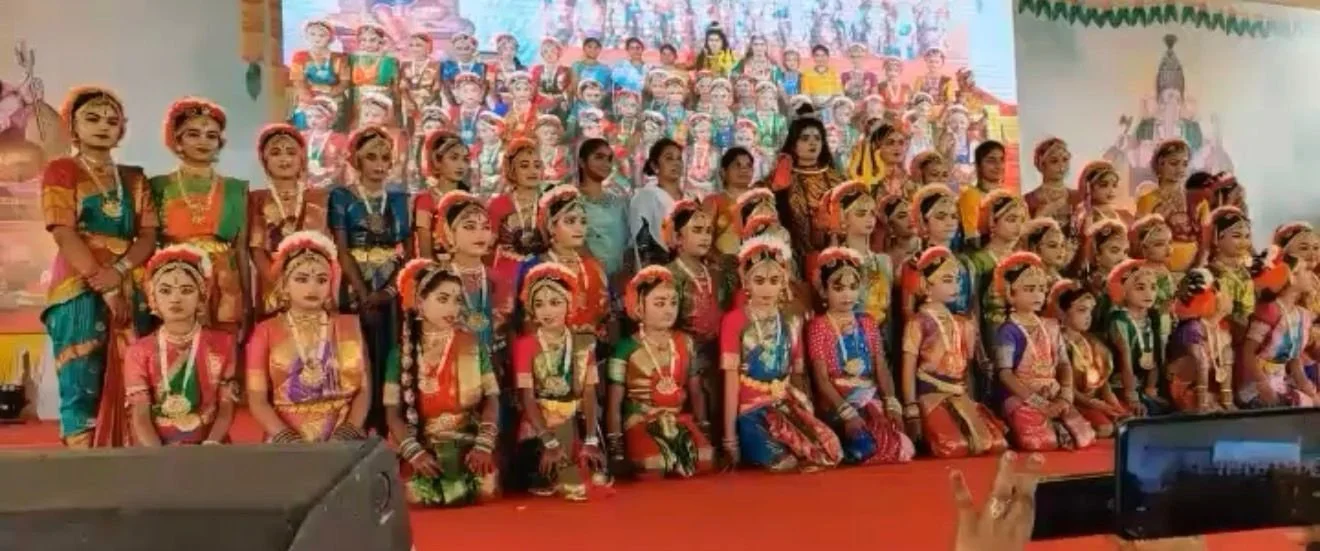 A large group of young children dressed in traditional colorful Indian costumes, posing on stage for a group photo during a cultural event or performance.