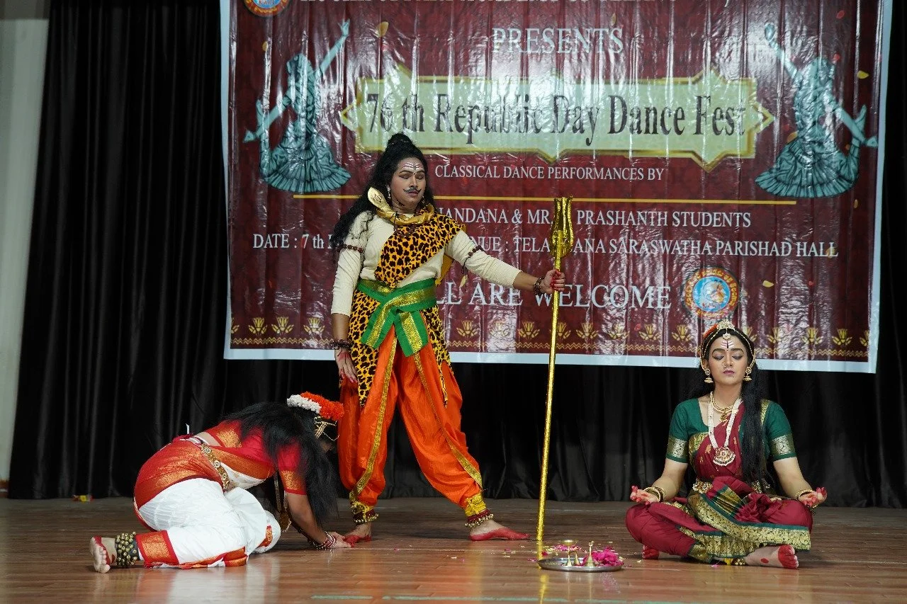 Three young women performing a classical dance on stage, dressed in traditional Indian costumes, with one woman standing holding a staff and two seated women on the floor.