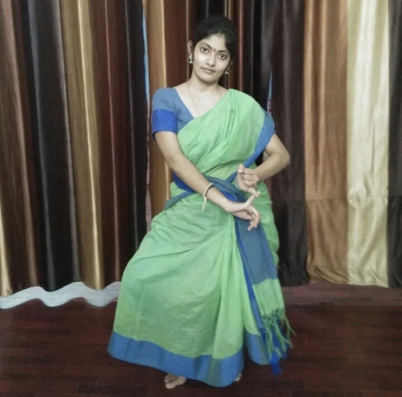 A woman in a green and blue sari standing in front of curtains.
