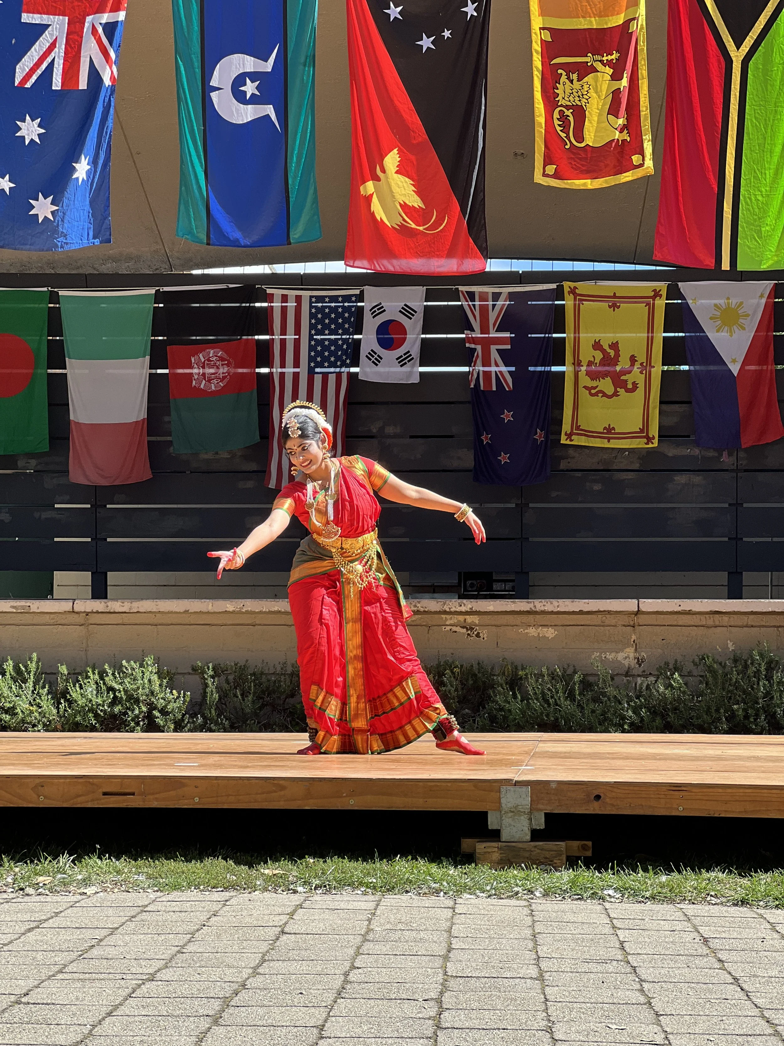 A dancer performing on an outdoor stage in traditional colorful attire, with multiple national flags hanging in the background.