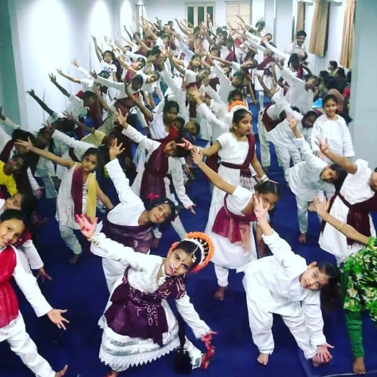 A group of children performing a dance in a classroom, dressed in white and maroon traditional Indian attire, with some wearing colorful accessories.