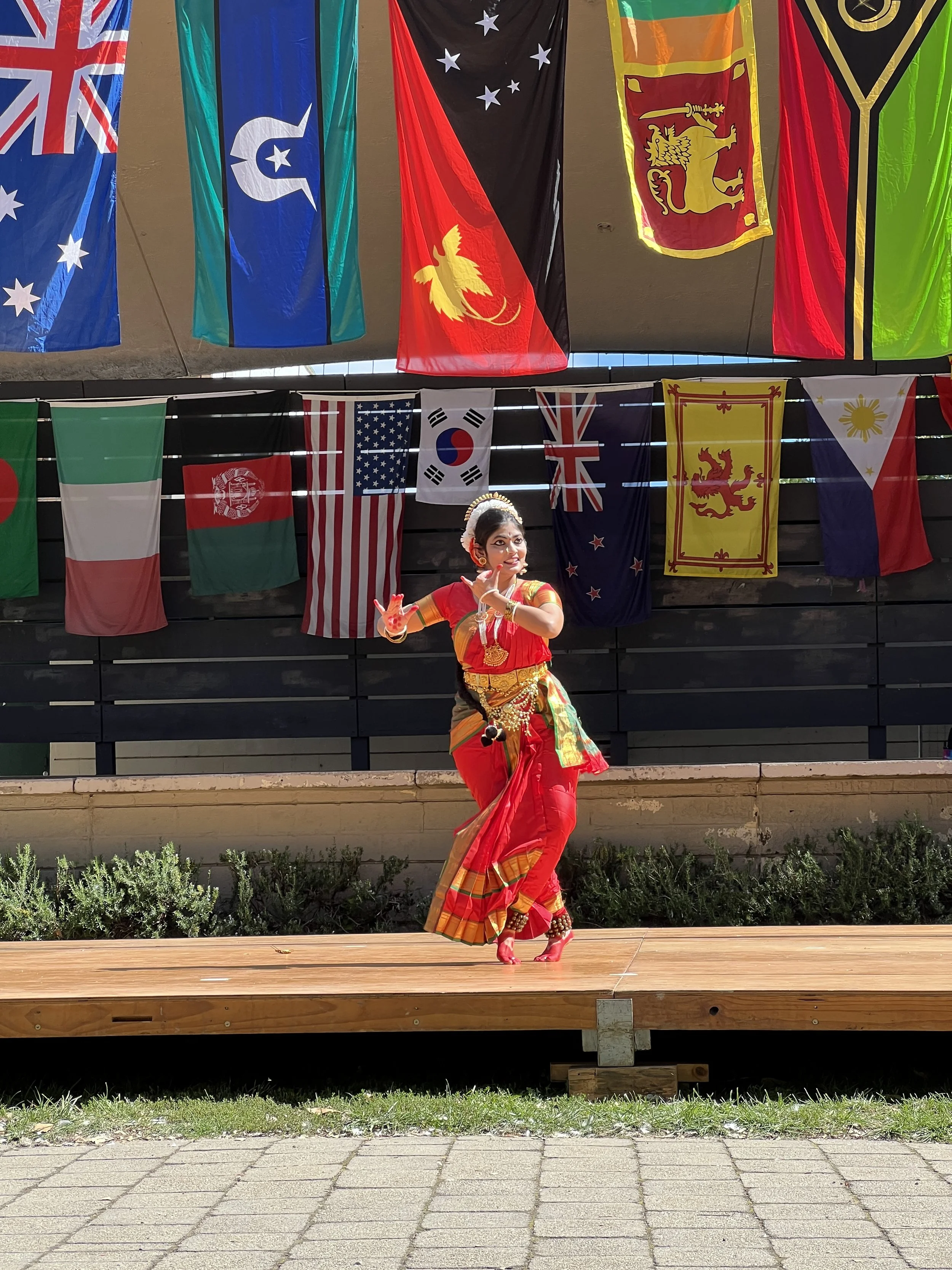 A woman dressed in traditional Indian dance attire performing on an outdoor stage with international flags hanging in the background.