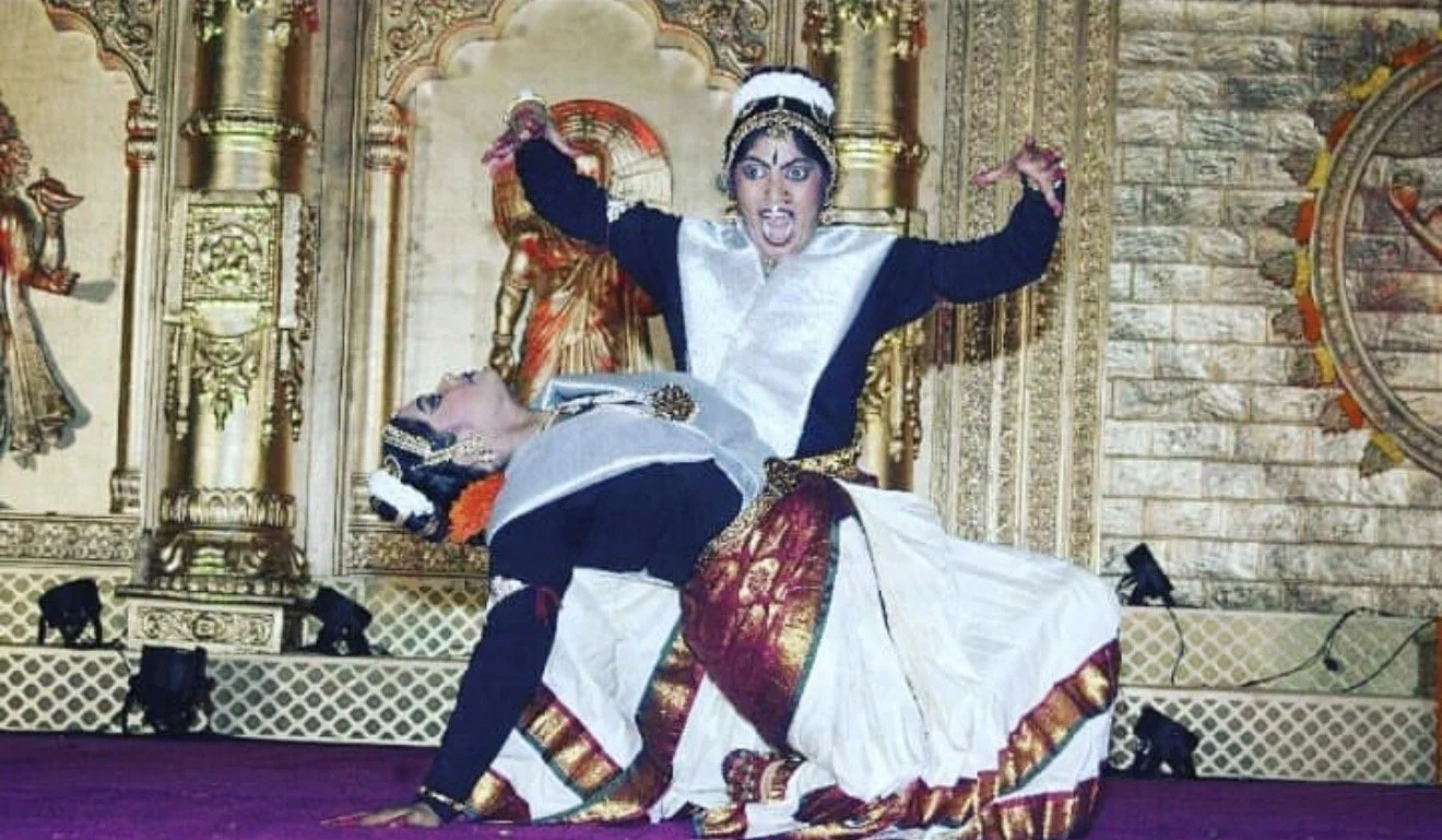 Two performers in traditional Indian dance costumes performing on stage, with ornate golden decorations in the background.