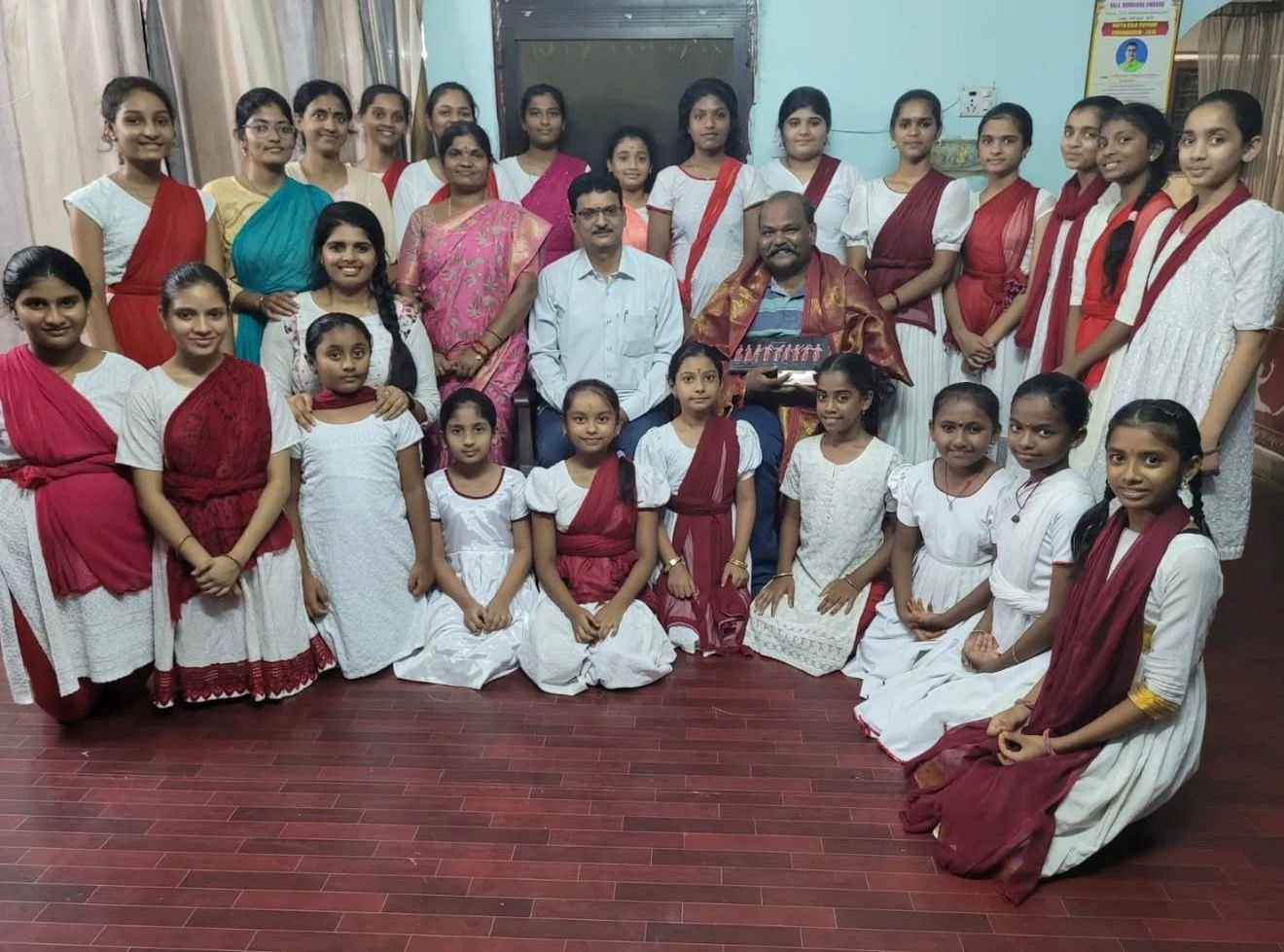 A group of girls and women in traditional Indian attire, along with two men, posing for a photo indoors. Some girls are seated on the floor, others are standing, in front of a dark window and light-colored walls.