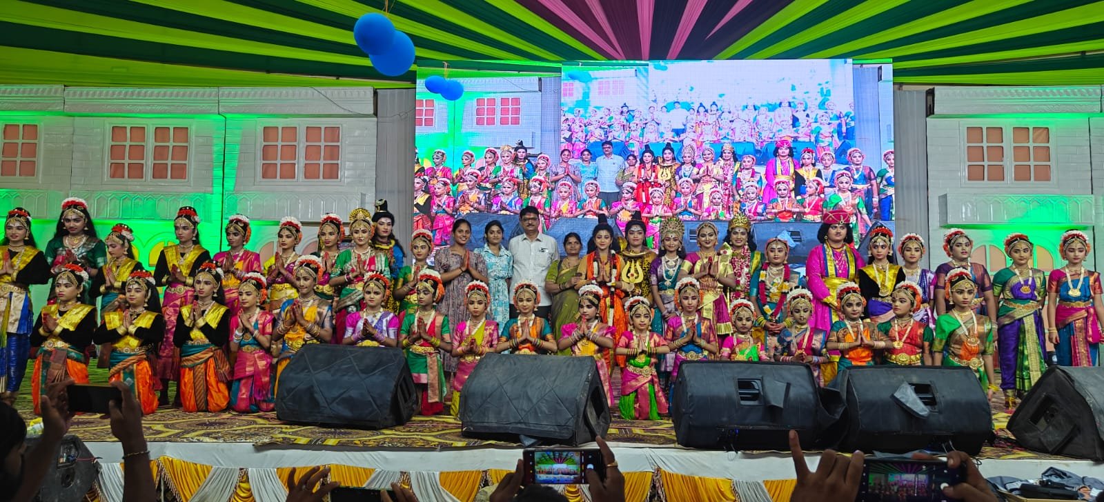 Children dressed in colorful traditional attire perform on stage during a cultural event, with adults standing behind them and a large screen projecting the event in the background.