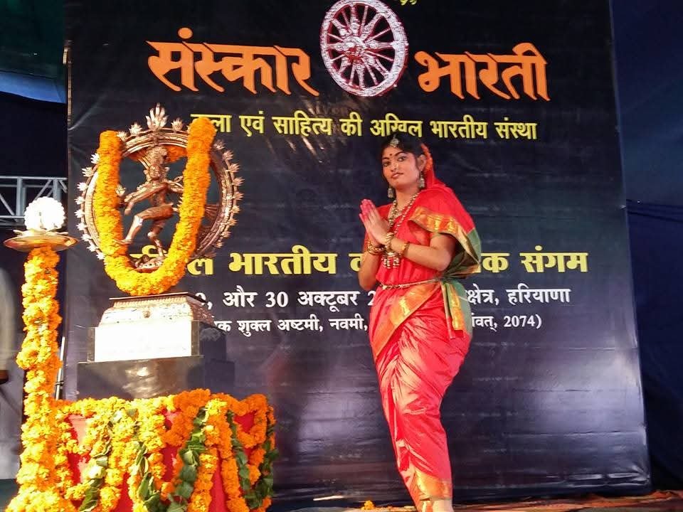 A woman dressed in traditional Indian attire standing next to a decorated statue of Lord Krishna during a celebration or festival, with a large banner in the background written in Hindi.