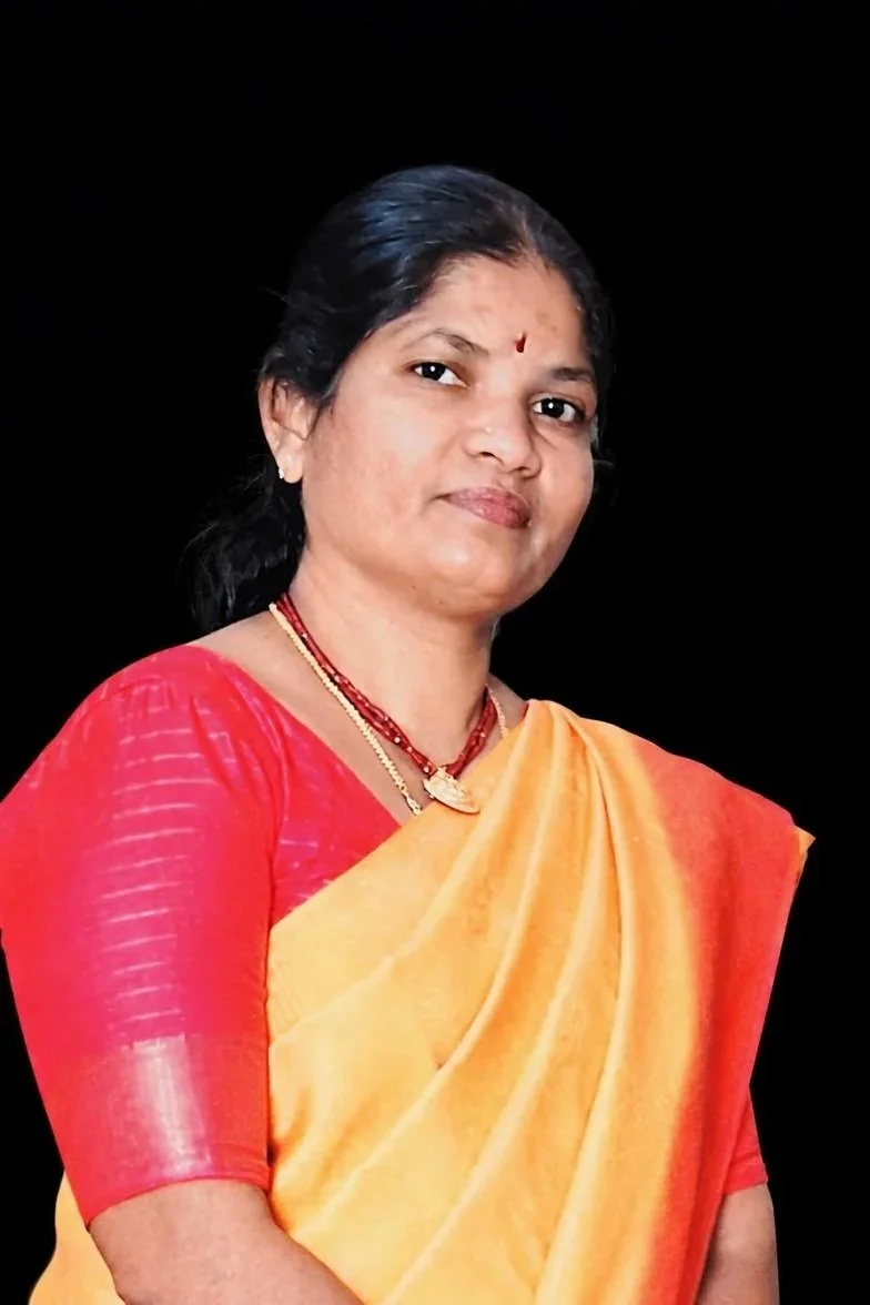 A woman wearing a yellow and orange saree with a red blouse, gold jewelry, and a small red bindi on her forehead, standing against a black background.
