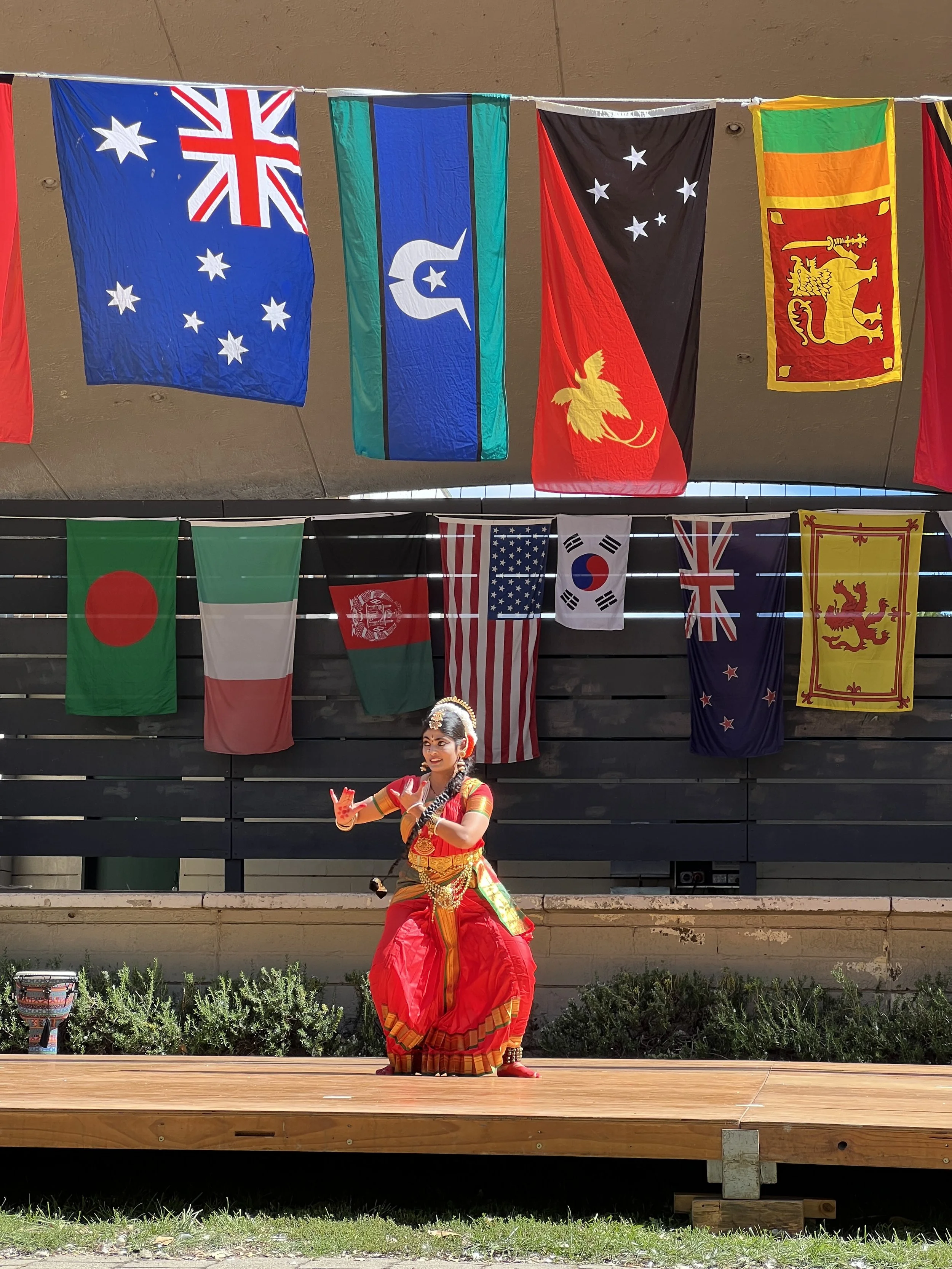 A young girl in traditional Indian dance attire performing on an outdoor stage with national flags hanging above her.
