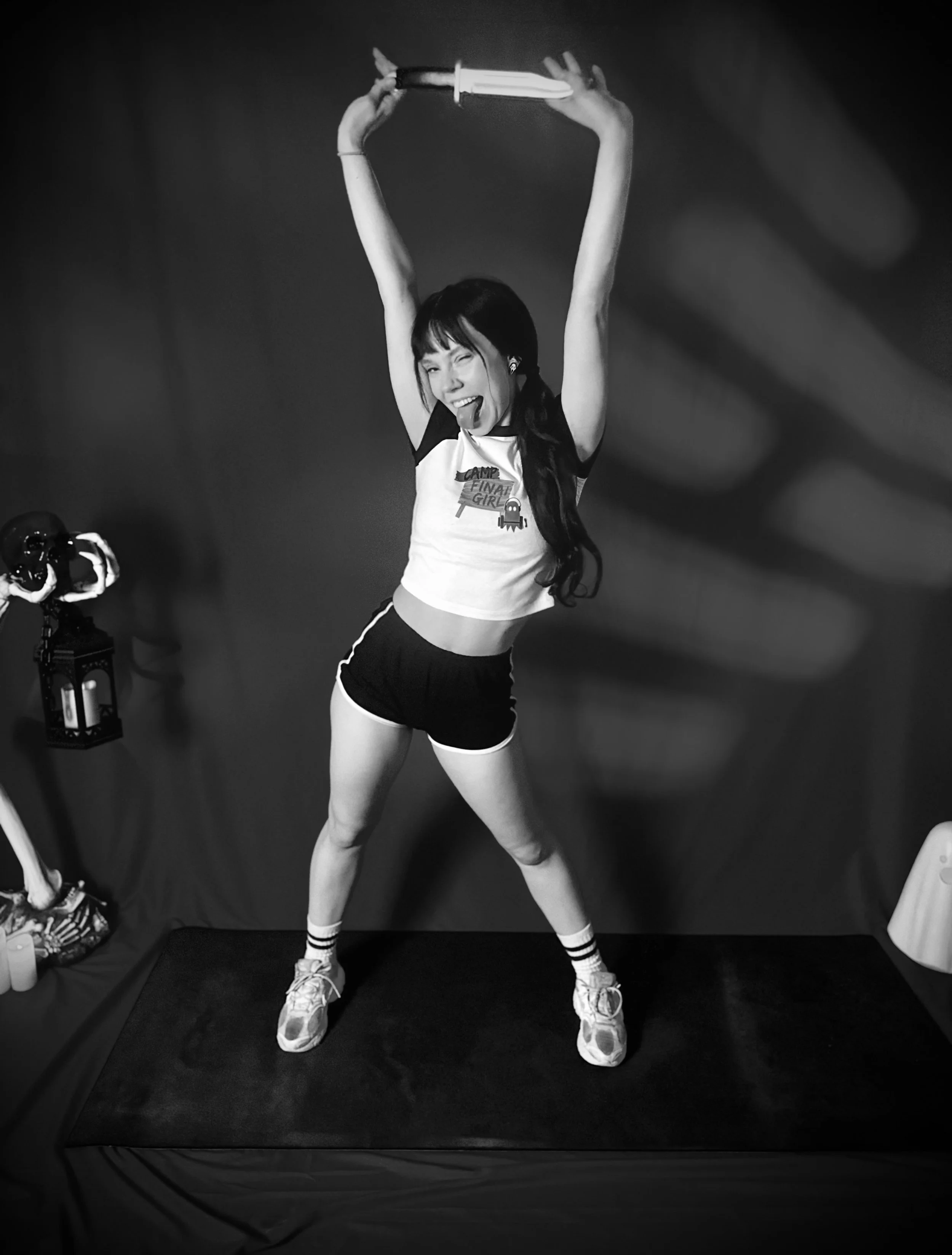 A young woman with long dark hair holding a large knife made of light above her head. She is smiling, sticking out her tongue, and wearing a T-shirt with a graphic that says 'Camp Final Girl.' She is in a casual athletic outfit with striped socks and sneakers, standing in a dark room with a lamp and camera nearby.