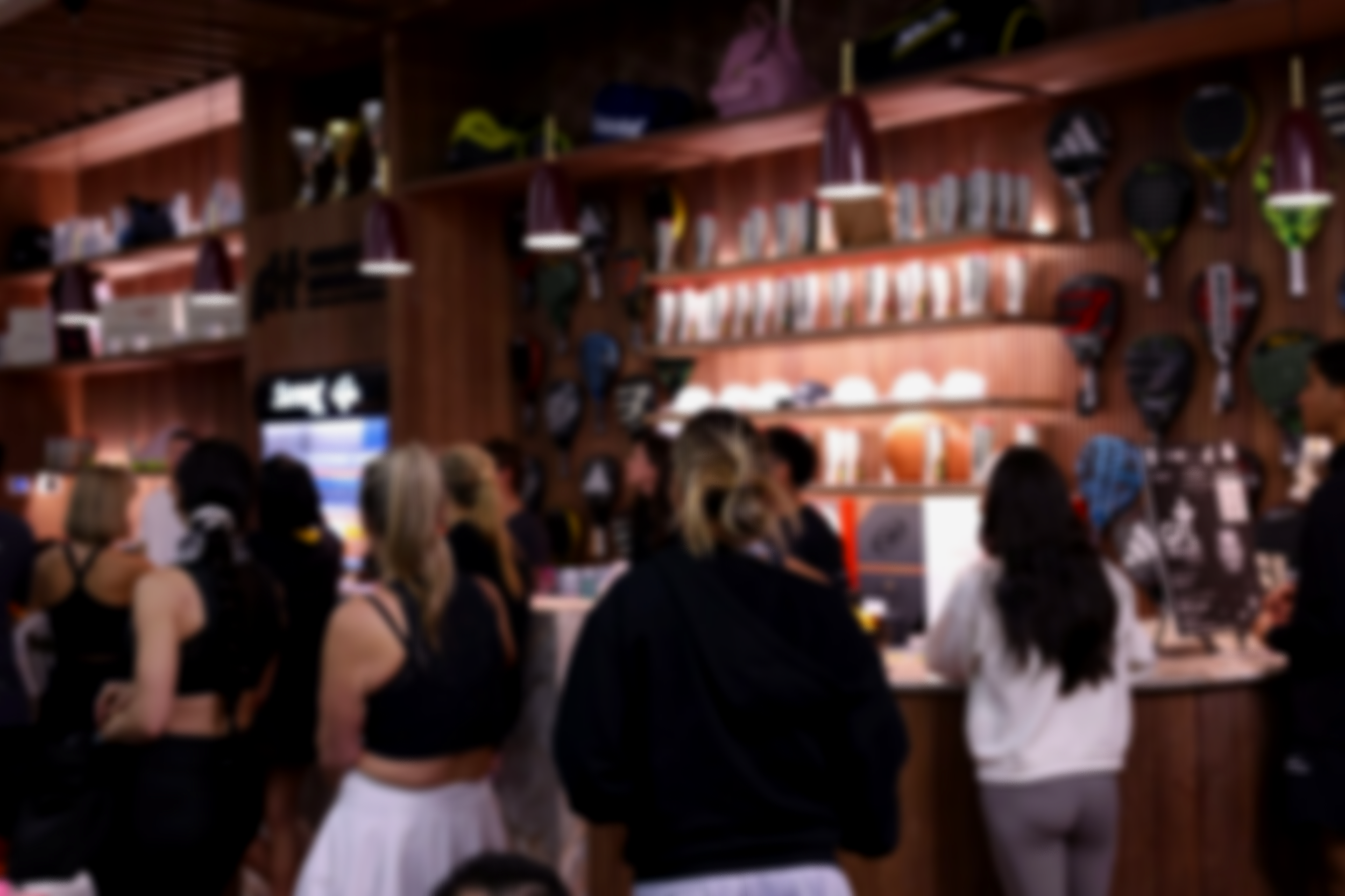People standing in an Indoor luxury padel club with padel racquets and balls on the wall behind them, decorated with shelves of balls and trophies.
