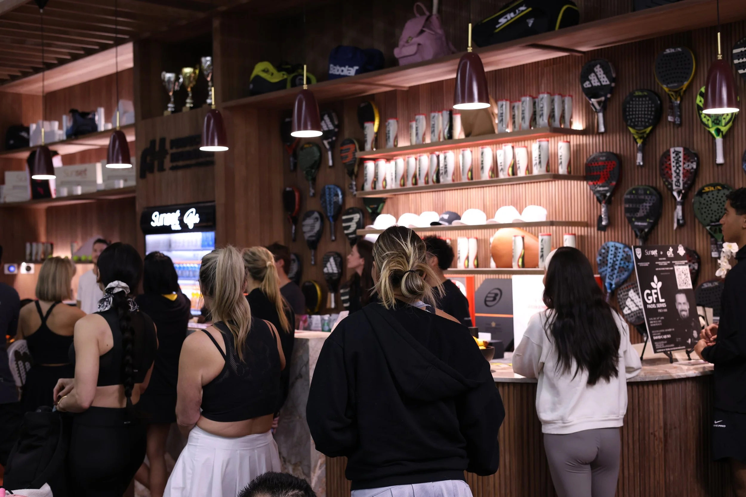 People standing in an Indoor luxury padel club with padel racquets and balls on the wall behind them, decorated with shelves of balls and trophies.