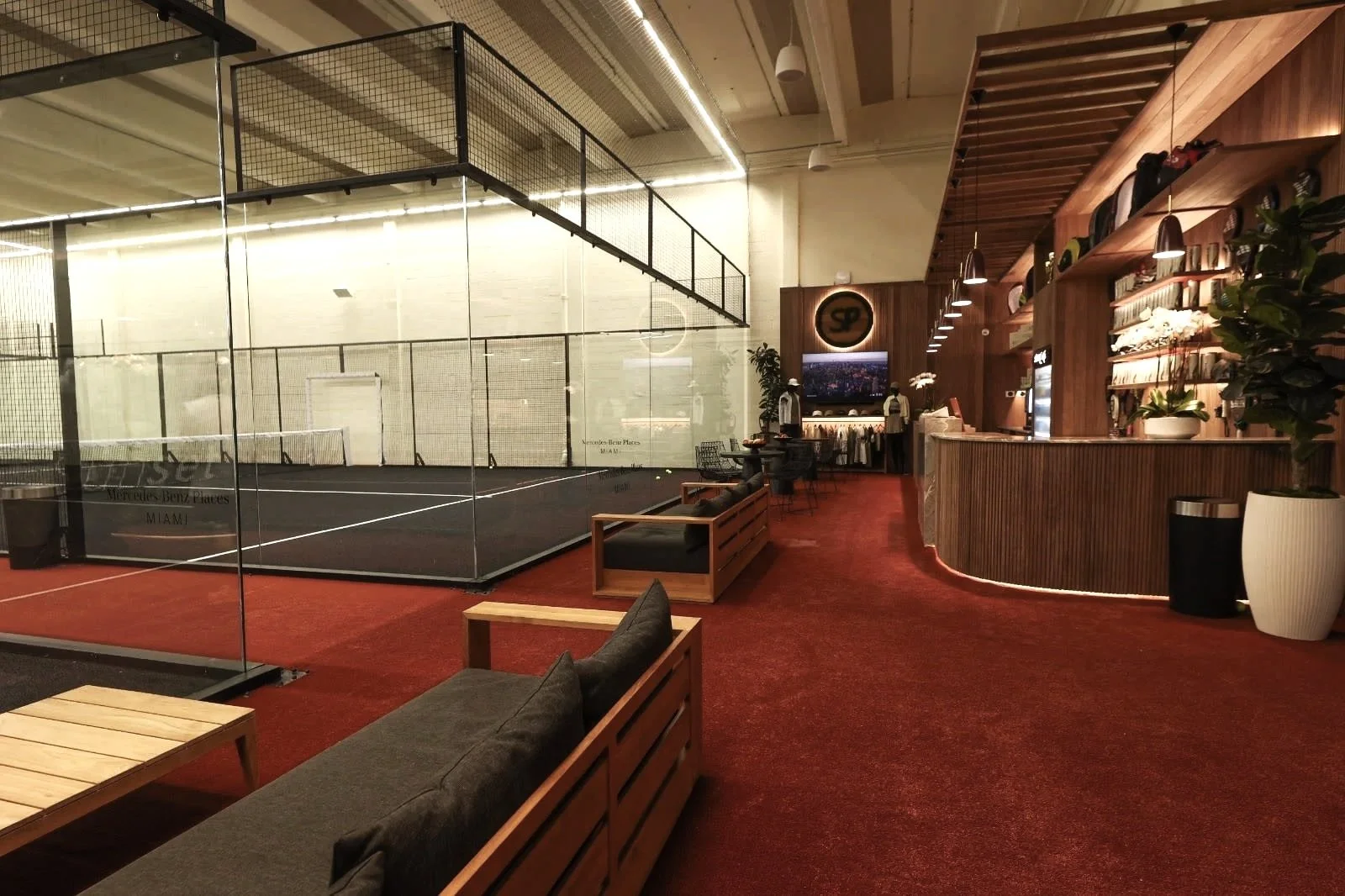 Indoor space with a padel court, adjacent to a lounge area with wooden furniture, couches, plants, and a counter with shelves, lights, and a television.