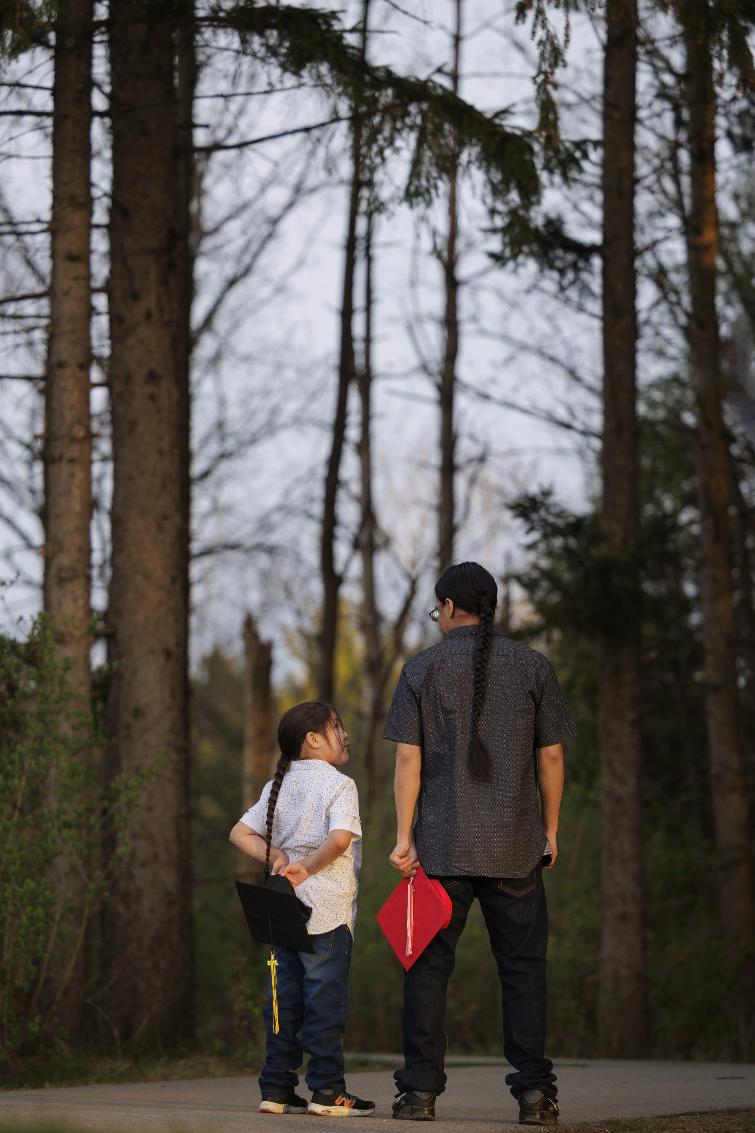 Kindergarten boy with long hair braided, holding black graduation cap looking up to a high school senior boy holding red graduation cap