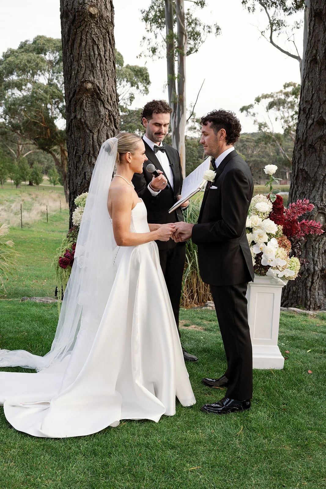 A couple getting married outdoors, holding hands, with a priest and officiant present. The bride is in a white wedding gown with a veil, and the groom in a black tuxedo. There are trees and a floral arrangement nearby.