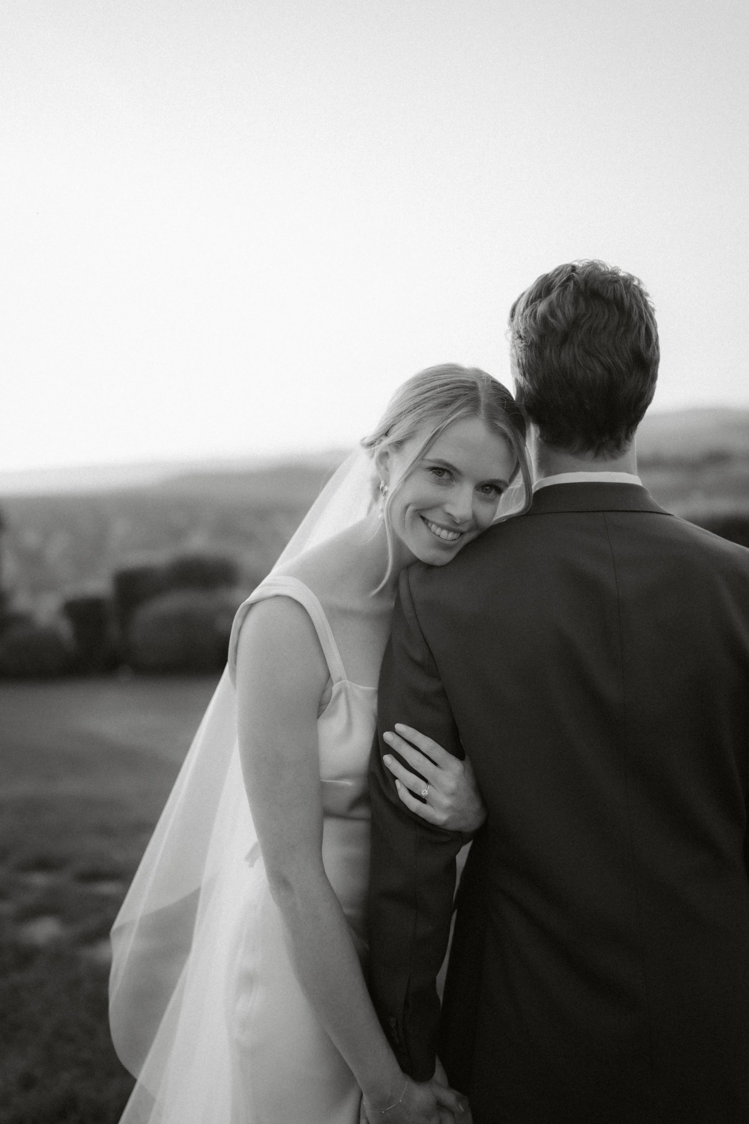 Black and white photo of a bride with blonde hair smiling and resting her head on a groom's shoulder. The bride's left hand shows a wedding ring. The groom, seen from the back, is wearing a dark suit, and they are outdoors with blurred landscape in the background.