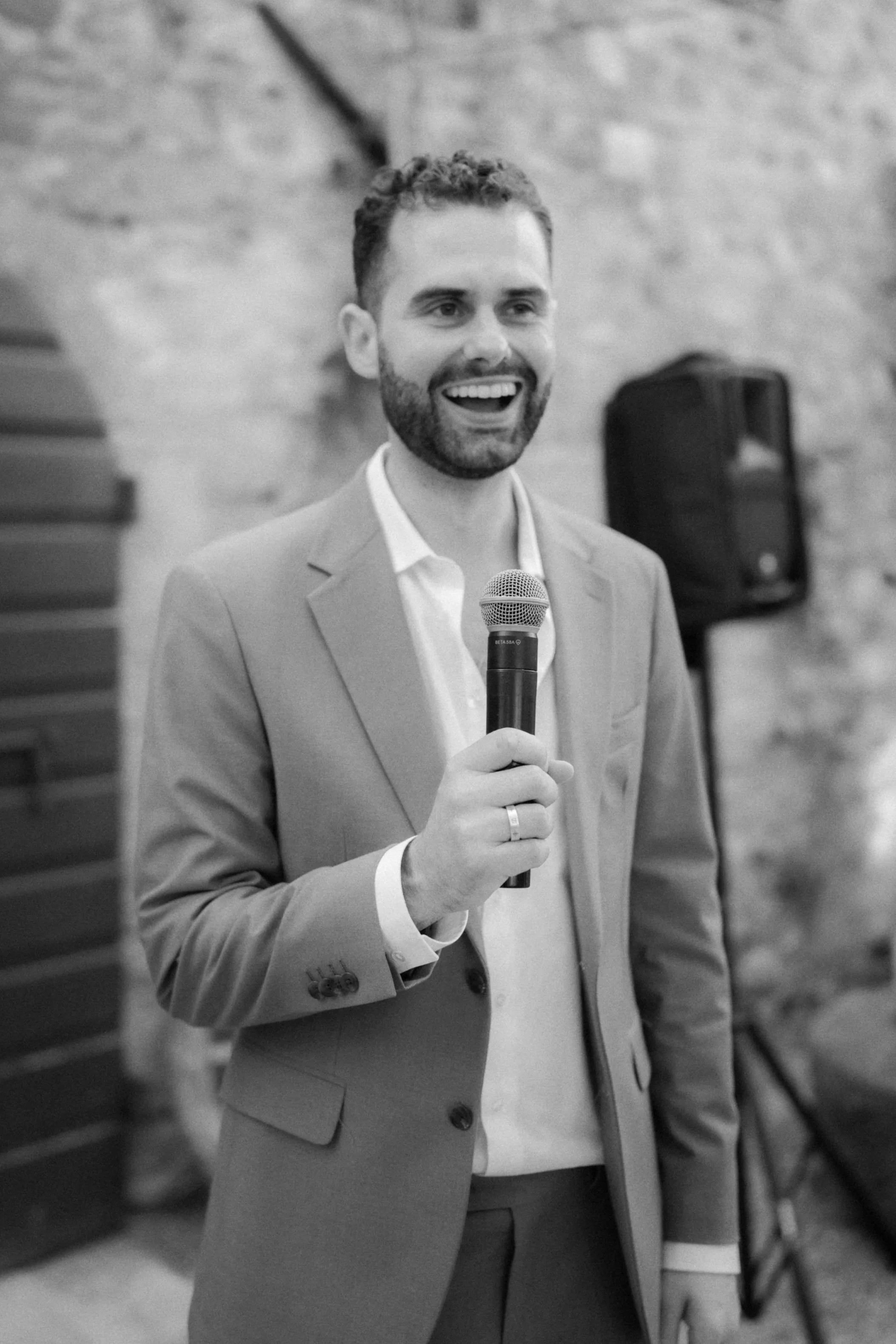 A man in a suit holding a microphone and speaking at an indoor event, with a brick wall and a speaker in the background.