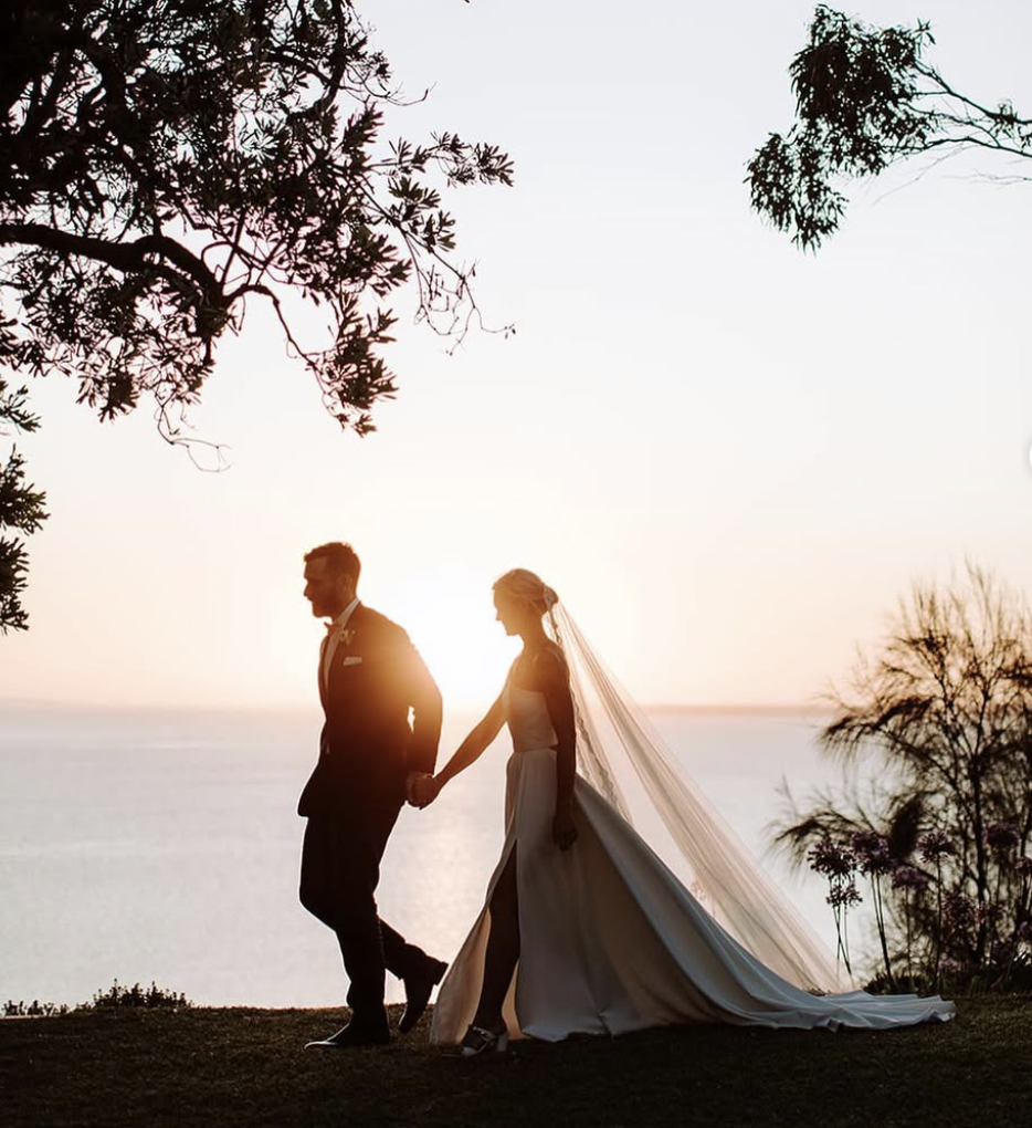 A silhouette of a bride and groom holding hands during sunset, near the water with trees in the background.