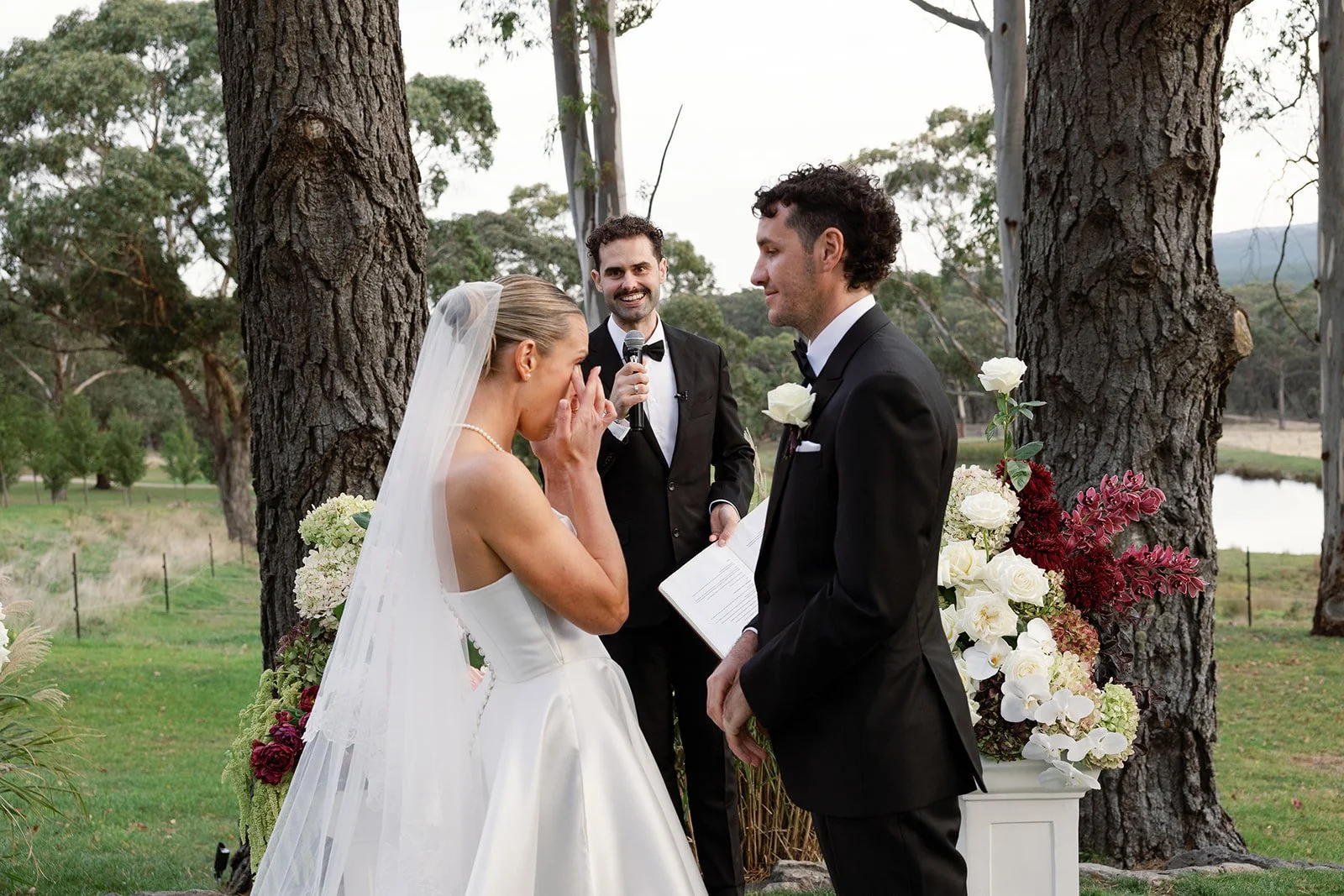 A bride and groom exchanging vows outdoors, with an officiant holding a microphone. The bride is wearing a white wedding gown with a veil, and the groom is in a black tuxedo with a white boutonniere. They are standing beside a floral arrangement near trees and a river.